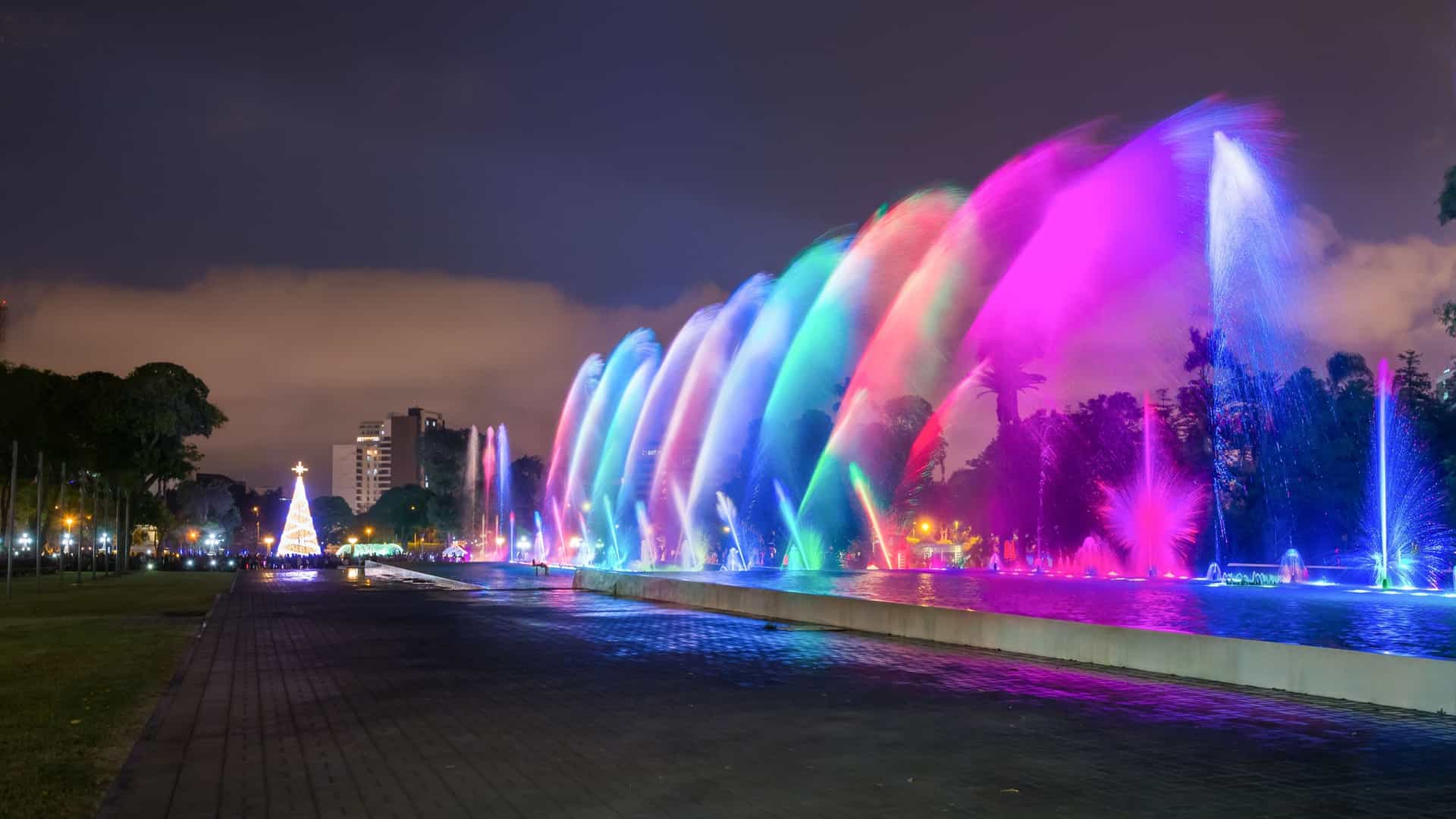 A nighttime photograph of the Magic Water Circuit in the Park of the Reserve (Parque de la Reserva) in Lima, Peru, showing a tall fountain jet illuminated in vibrant colors like blue and pink, with light reflections visible in the surrounding water.