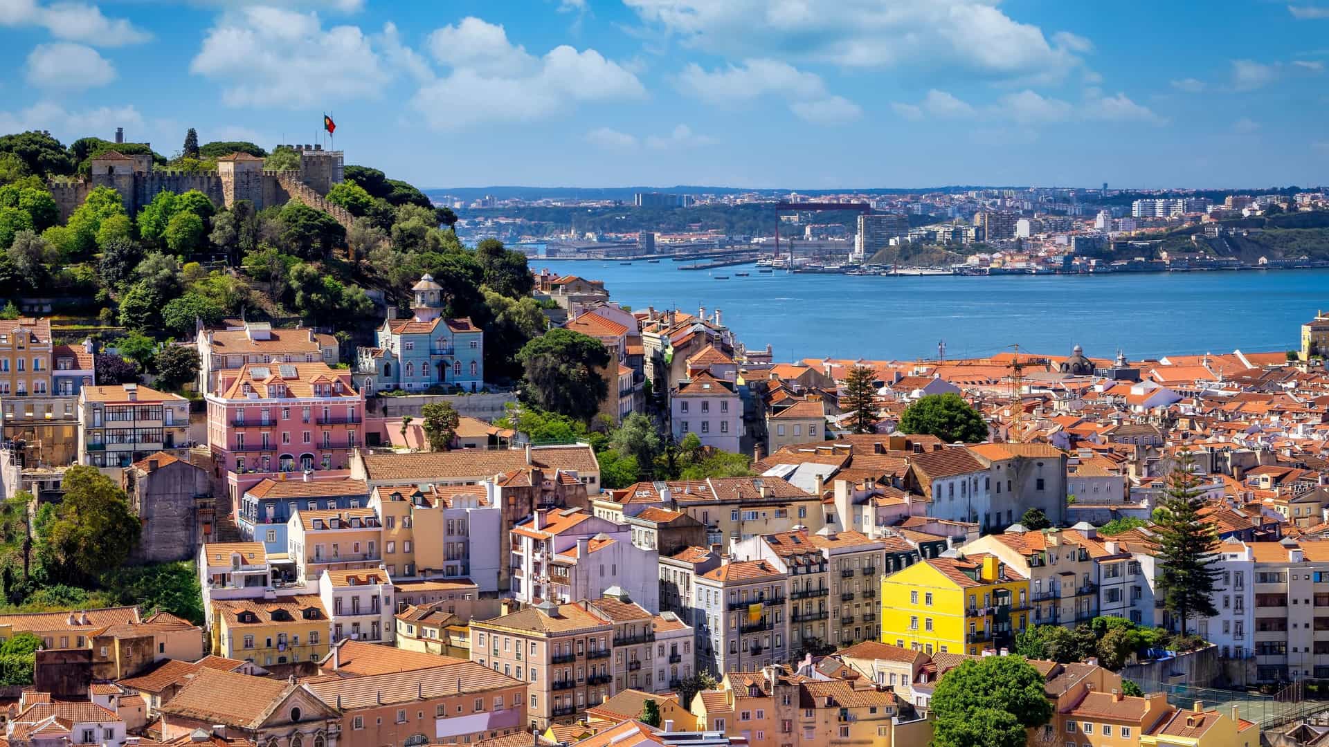  A cityscape of Lisbon, Portugal, seen from a high vantage point at Castelo de São Jorge (St. George's Castle). The image captures a dense urban landscape of buildings with traditional red tiled rooftops, with the Tagus River visible in the background.
