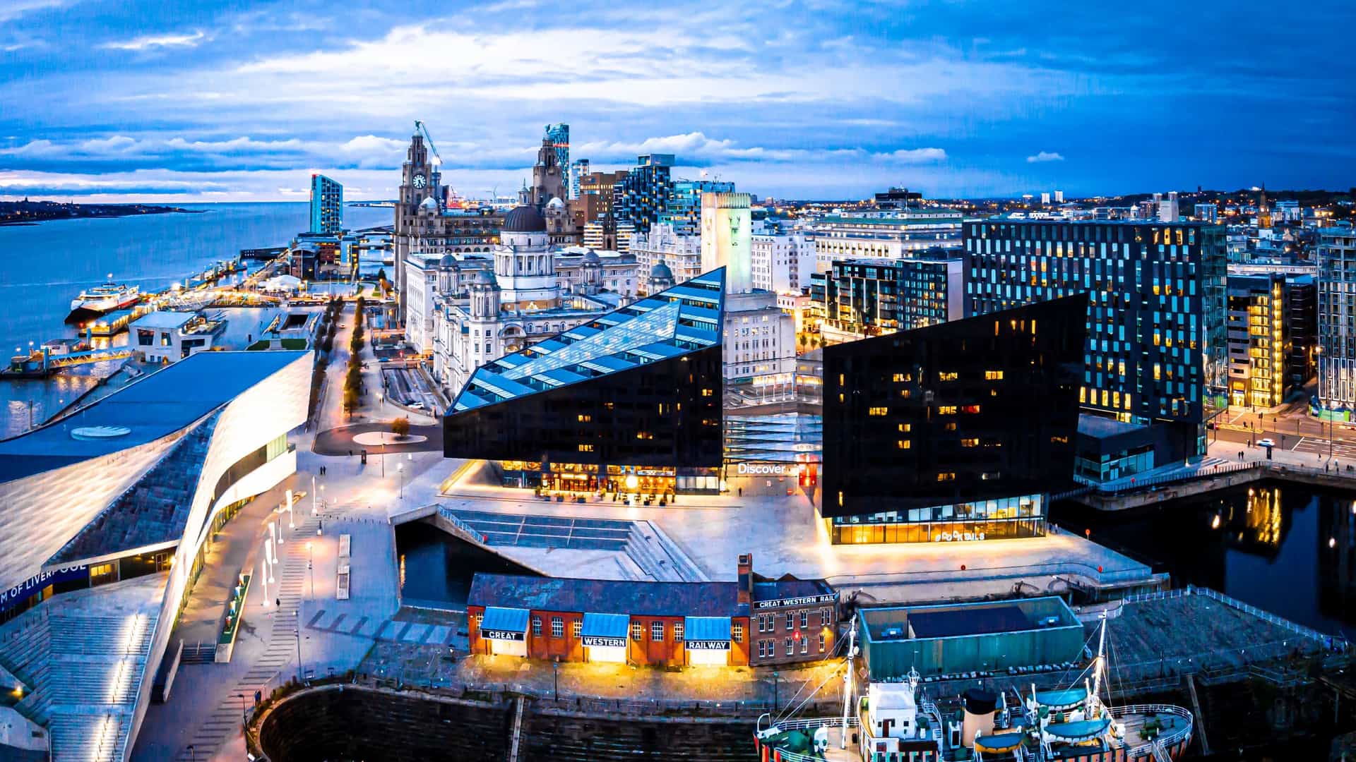  An aerial view of the Royal Albert Dock in Liverpool, showing the square dock basin surrounded by the iconic red brick warehouses. The River Mersey is visible in the background, along with the broader cityscape of Liverpool.