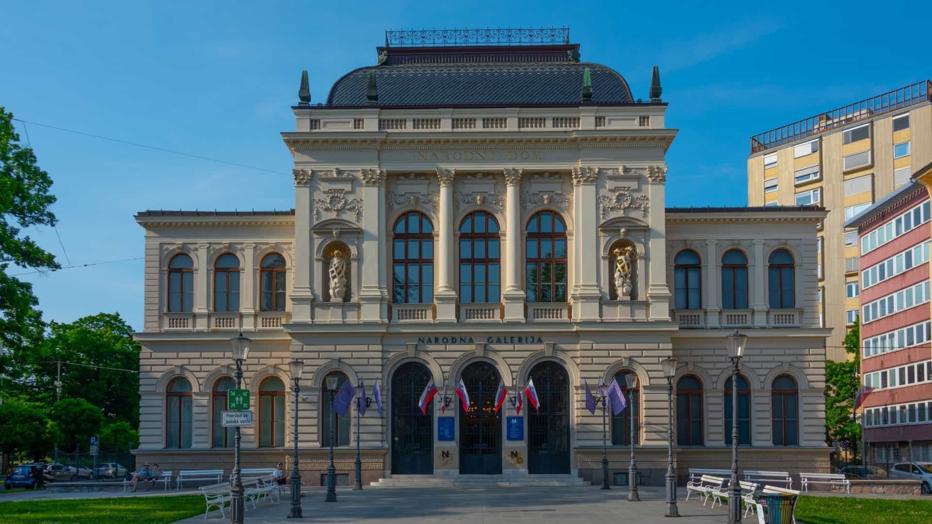 A view of the National Gallery of Slovenia building in Ljubljana. The gallery is housed in a grand, historic building, likely in a neo-Renaissance or similar classical style, with a striking yellow facade. The image may show the building's ornate architecture, including columns or detailed ornamentation, and is set against a daytime sky, typical of a cultural landmark photo.