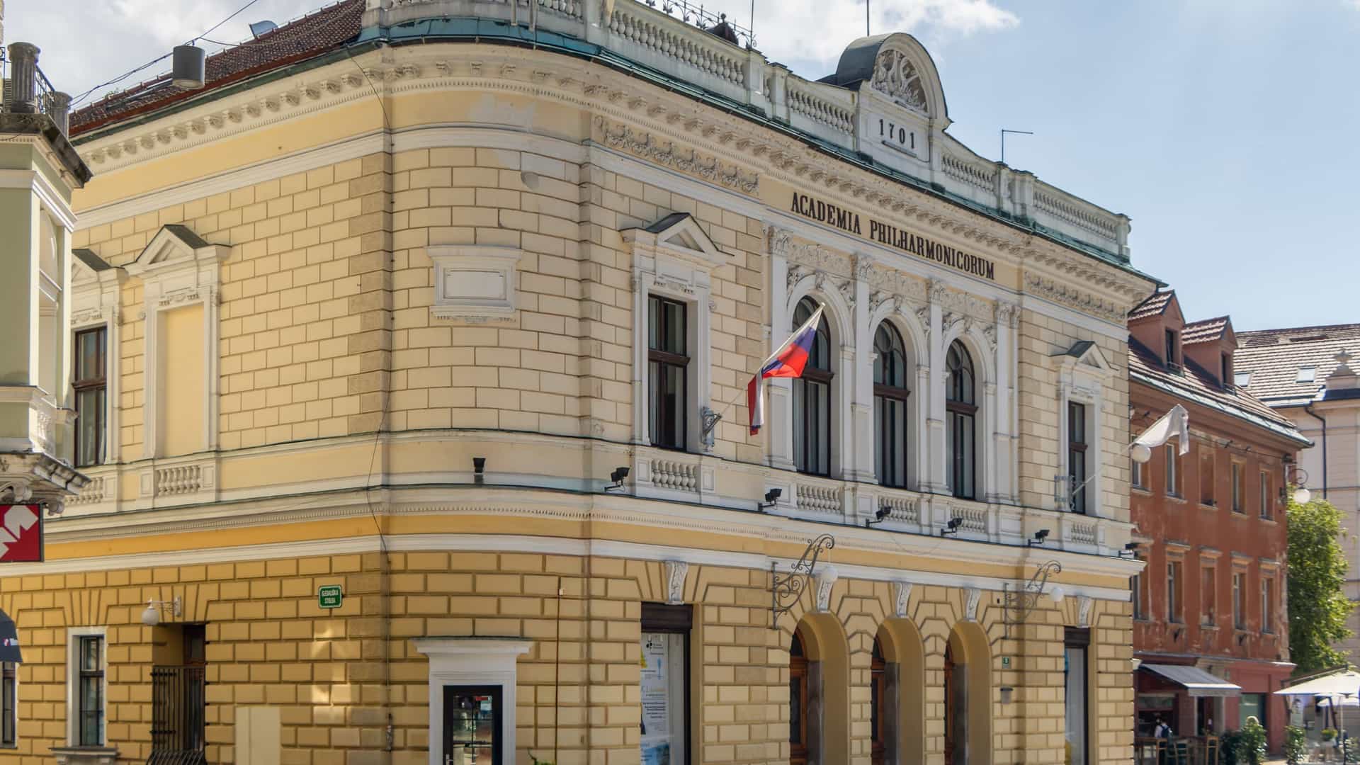 A photograph capturing the Neo-Renaissance facade of the Slovenian Philharmonic Building (Academia Philharmonicorum) in Ljubljana, Slovenia. The elegant, light-colored, historic building is shown from the street level on Congress Square. The building's architecture is characteristic of the time it was constructed (1891), featuring rounded corners. The name "Academia Philharmonicorum" is typically visible in relief lettering across the upper portion of the facade.