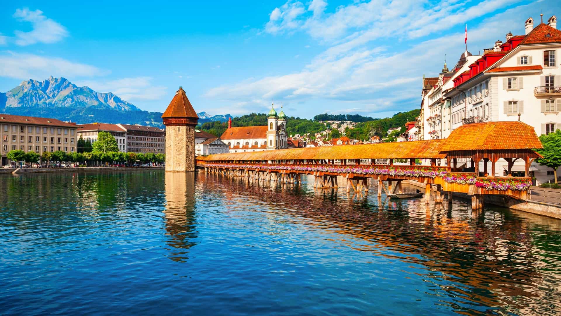  A photograph of the Kapellbrücke (Chapel Bridge), a covered wooden footbridge, spanning the Reuss River in Lucerne, Switzerland. The bridge is shown alongside the octagonal Wasserturm (Water Tower). The cityscape of Lucerne is visible in the background, with the Alps faintly outlined against the sky.