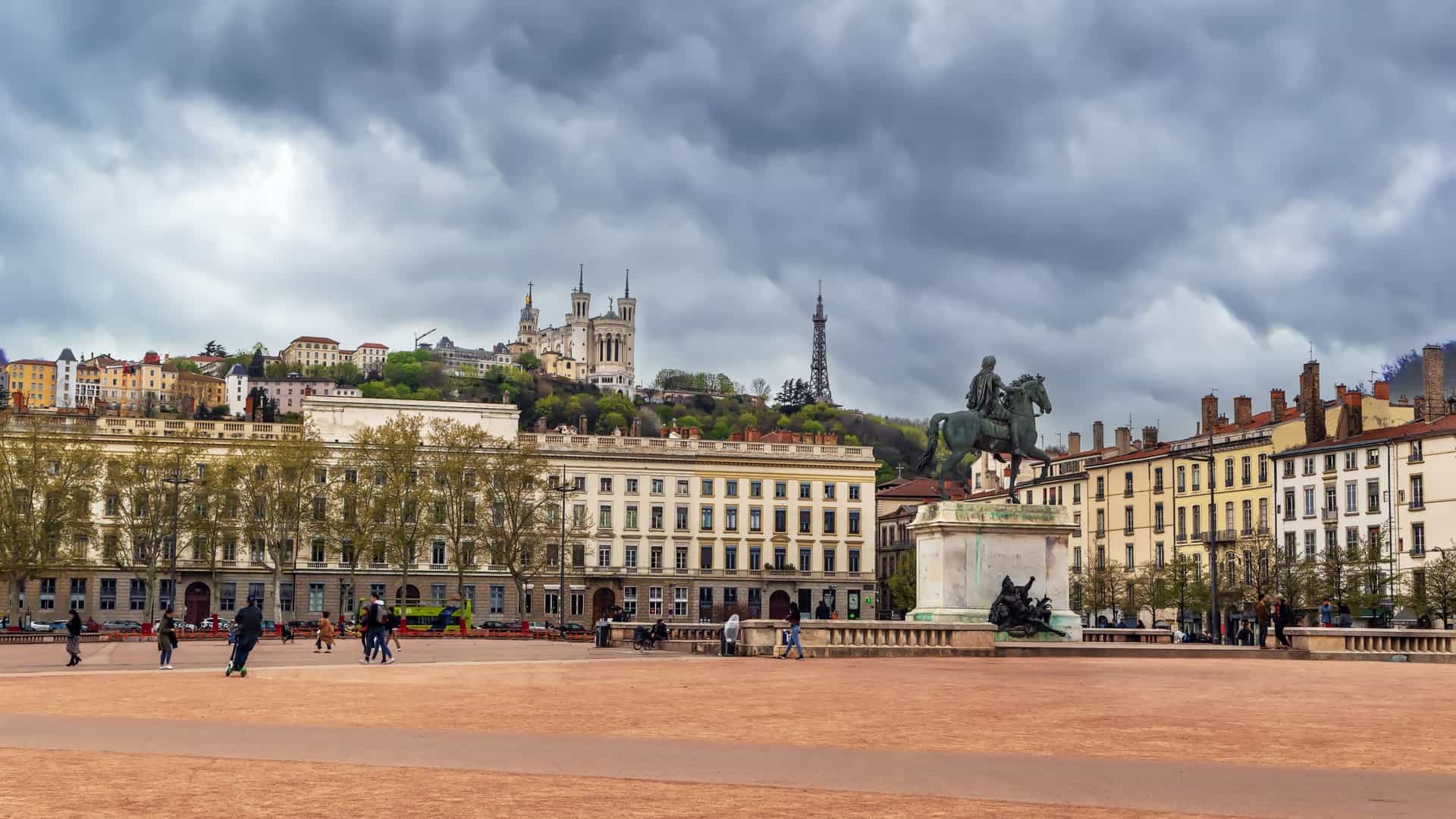 An expansive view of Place Bellecour in Lyon, France, one of the largest pedestrian squares in Europe. The image typically captures the large, open, unpaved square with the bronze equestrian statue of King Louis XIV prominently featured in the center. The backdrop includes the uniformly structured, historic buildings that frame the square, and often the Notre-Dame de Fourvière Basilica is visible on the hill in the distance.