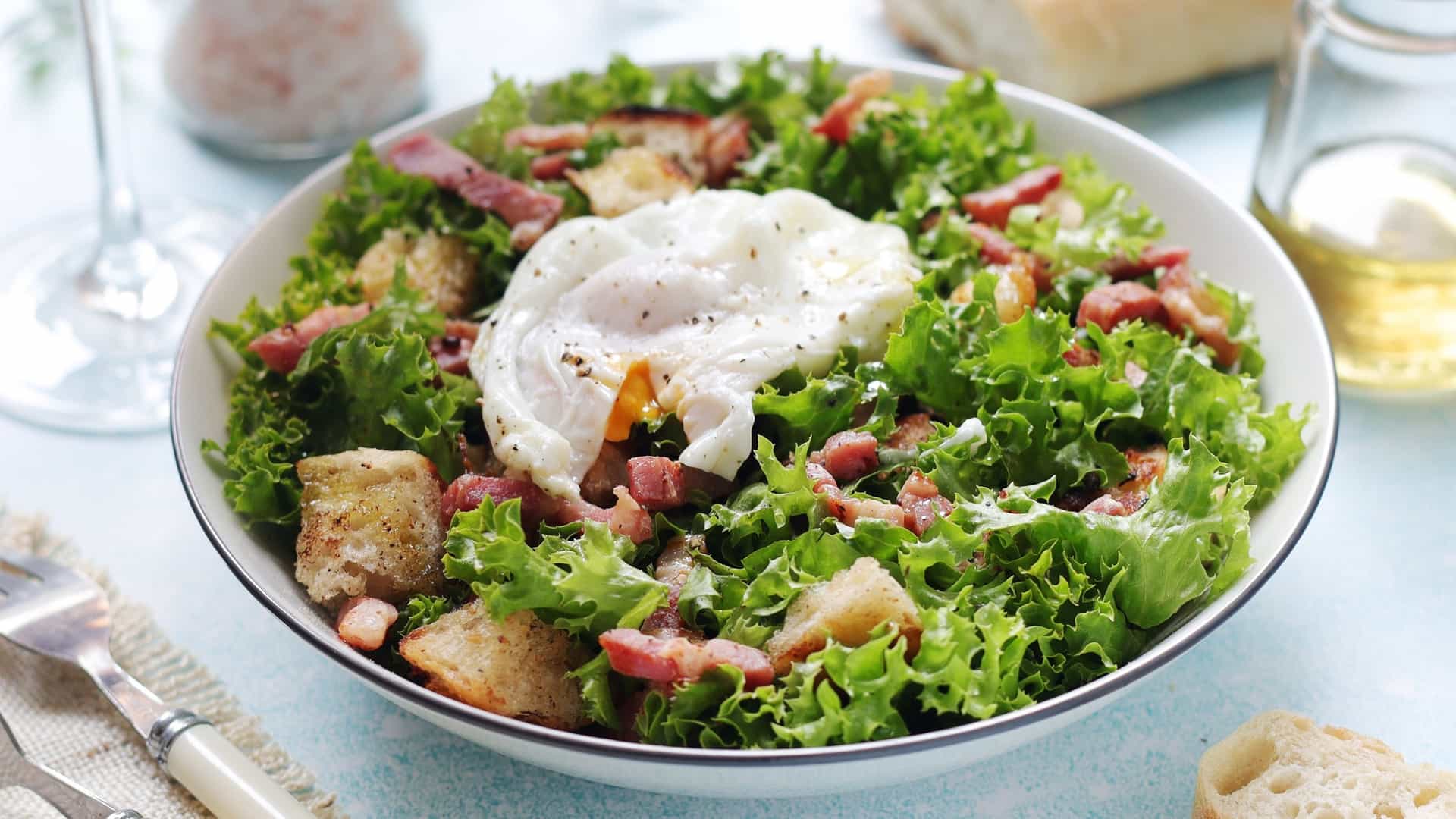 A close-up, top-down view of a bowl of Salad Lyonnaise served for lunch. The salad is composed of frisée lettuce (a curly, bitter green) tossed in a warm vinaigrette, and generously topped with crispy, thick-cut bacon pieces (known as lardons), and often a golden-yolked poached egg and croutons.