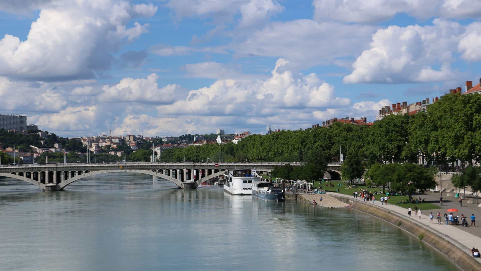 A sunny, panoramic view of the Rhône River as it flows through the city of Lyon, France. The image captures the modern development of the Berges du Rhône (Banks of the Rhône) on the left bank, which includes a wide, paved promenade, greenery, and trees. The river itself is lined with boats or barges, and the colorful, historic residential buildings of Lyon are visible on the opposite bank or across one of the bridges. The view highlights the urban leisure space created along the river.