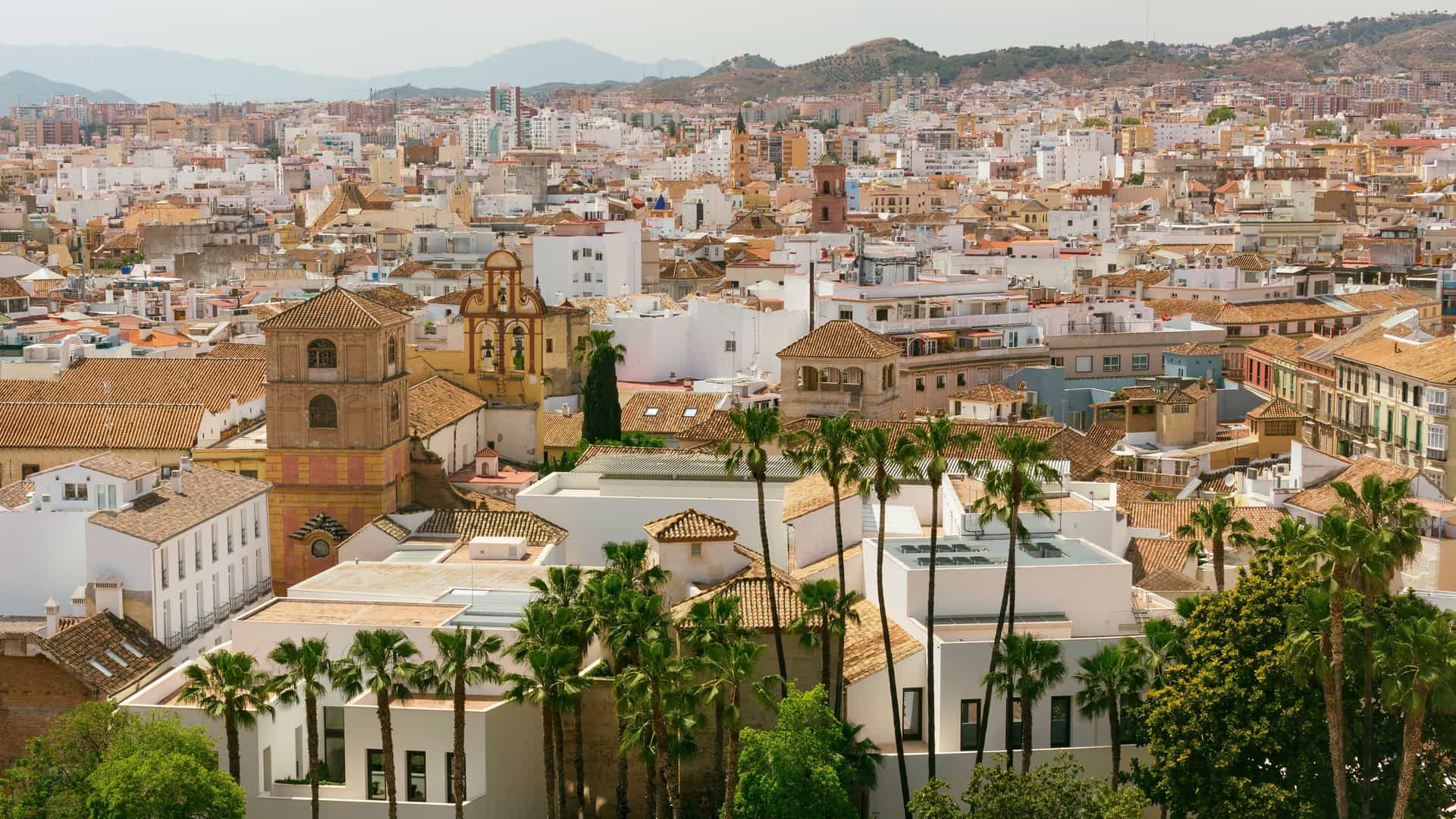 Panoramic view of the Picasso Museum (Palacio de Buenavista) in Malaga. The image features the museum's historic, tiled rooftop and architecture in the foreground, with the dense, white buildings and urban landscape of the city of Malaga stretching out towards the distant horizon.