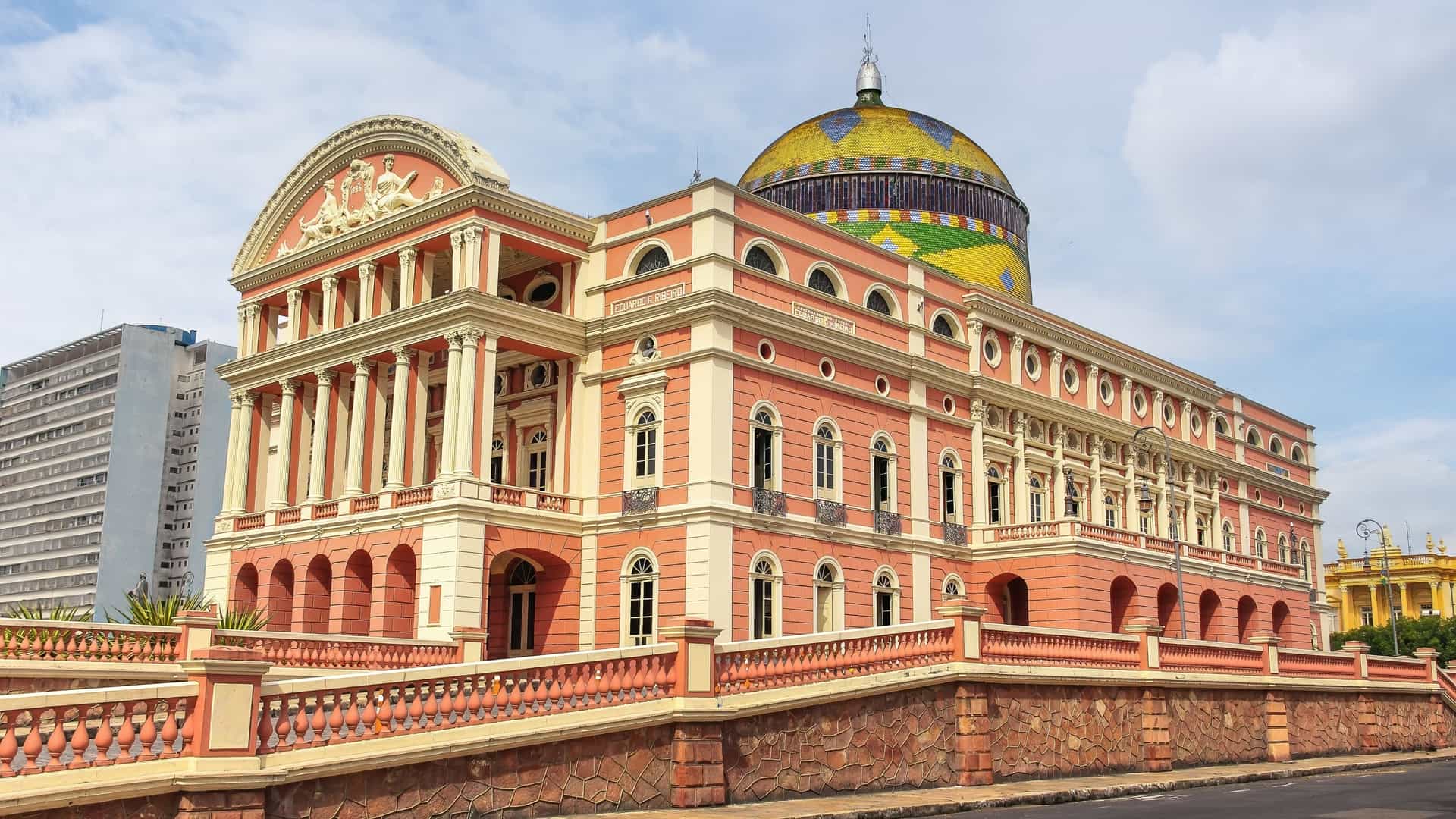 An exterior photo of the Teatro Amazonas (Amazonas Theatre), an opulent, pink Renaissance-style opera house with a distinctive domed roof tiled in the colors of the Brazilian flag (green, yellow, and blue), located in Manaus, Amazonas, Brazil.