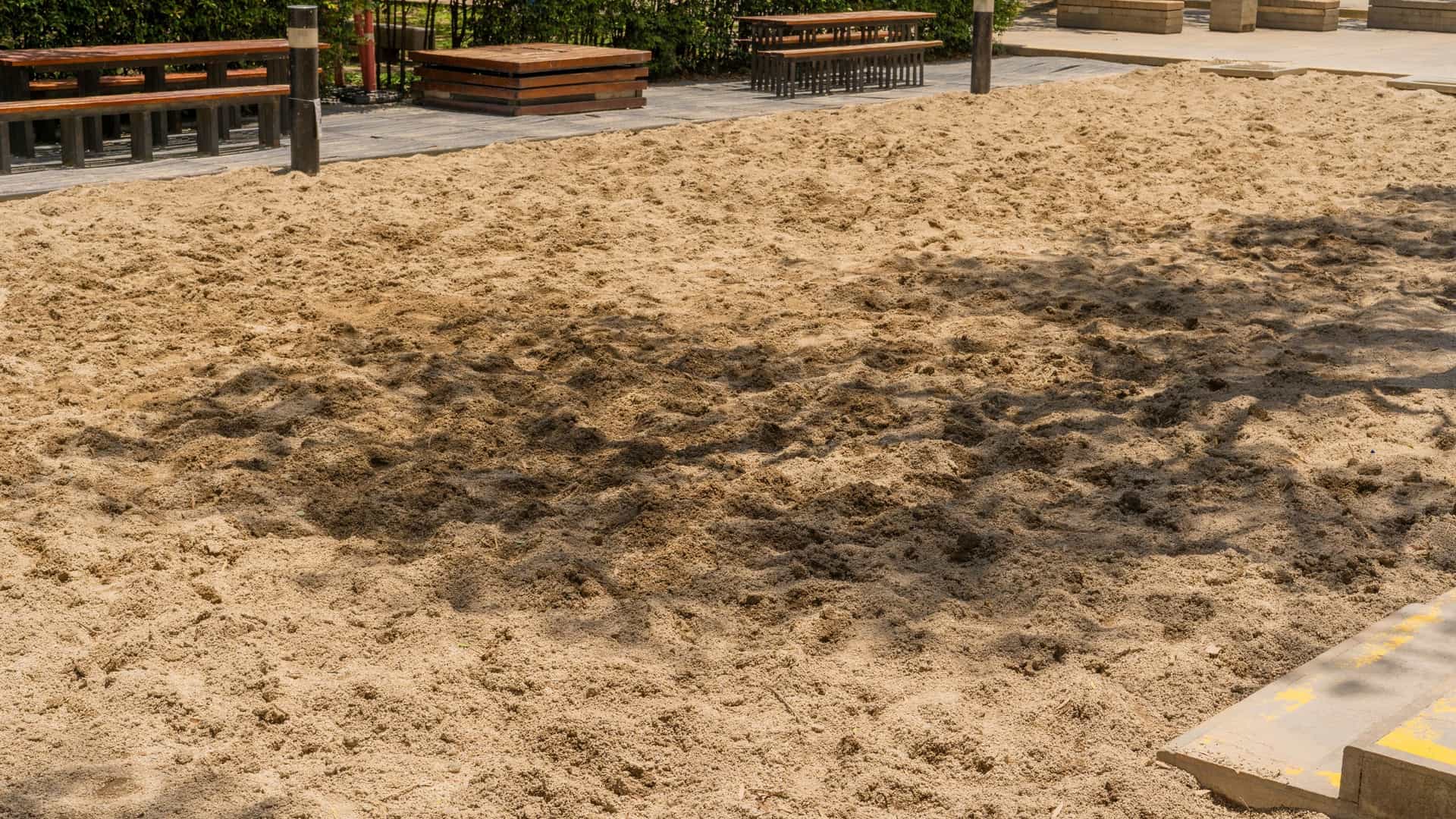 The large, excavated sandpit area of the Parque de los Pies Descalzos (Barefoot Park) in Medellín, Colombia. The loose, light brown sand covers the foreground, with seating areas and the surrounding hedges visible in the background. The park is designed for visitors to walk barefoot and engage with the environment.