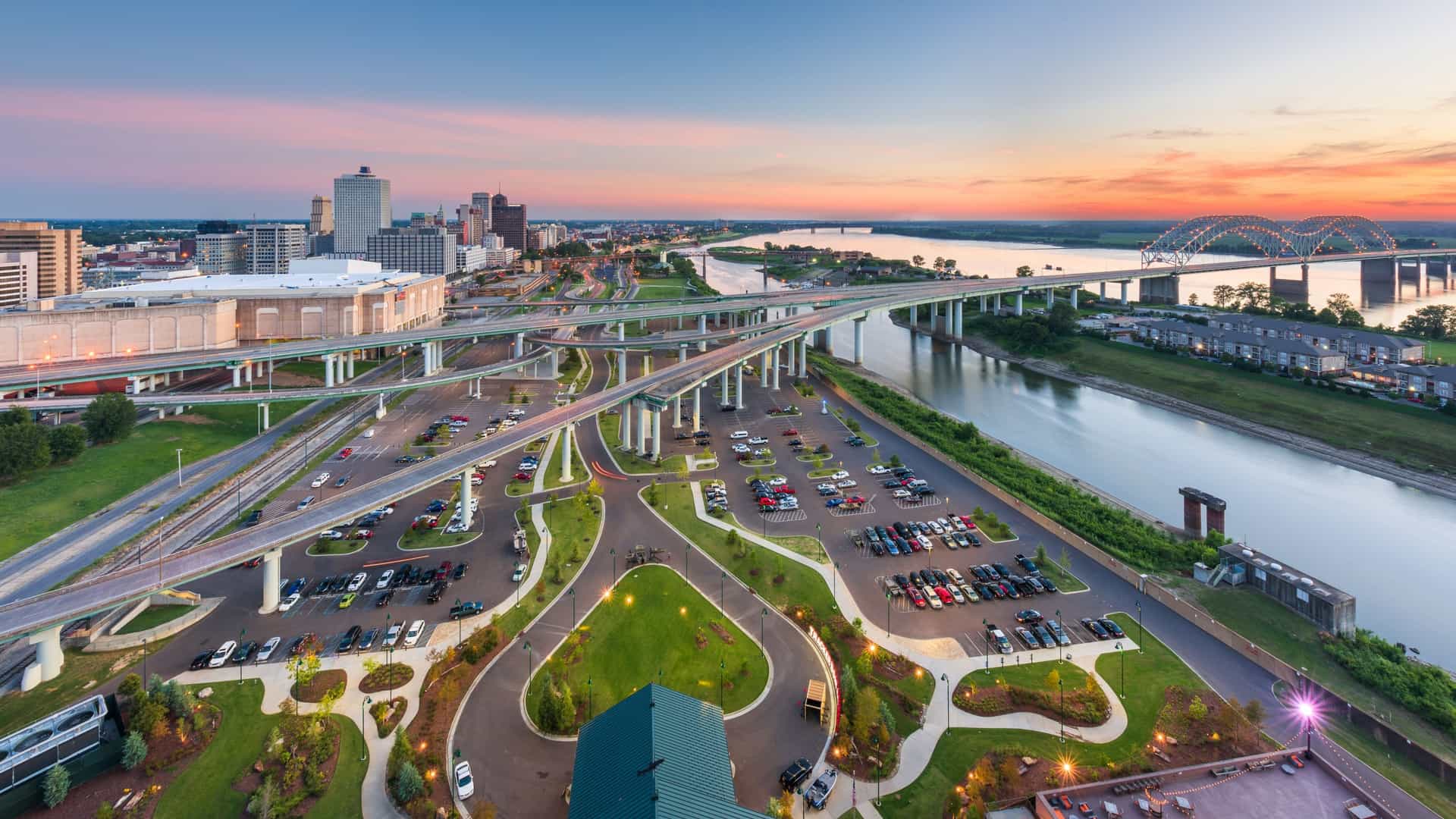  An aerial view of the Memphis, Tennessee, skyline at dusk. The image captures the downtown area and the Mississippi River, with the iconic Hernando de Soto Bridge, which carries Interstate 40, visible spanning the river. The city lights are beginning to illuminate the urban landscape, creating a dramatic contrast with the setting sun.