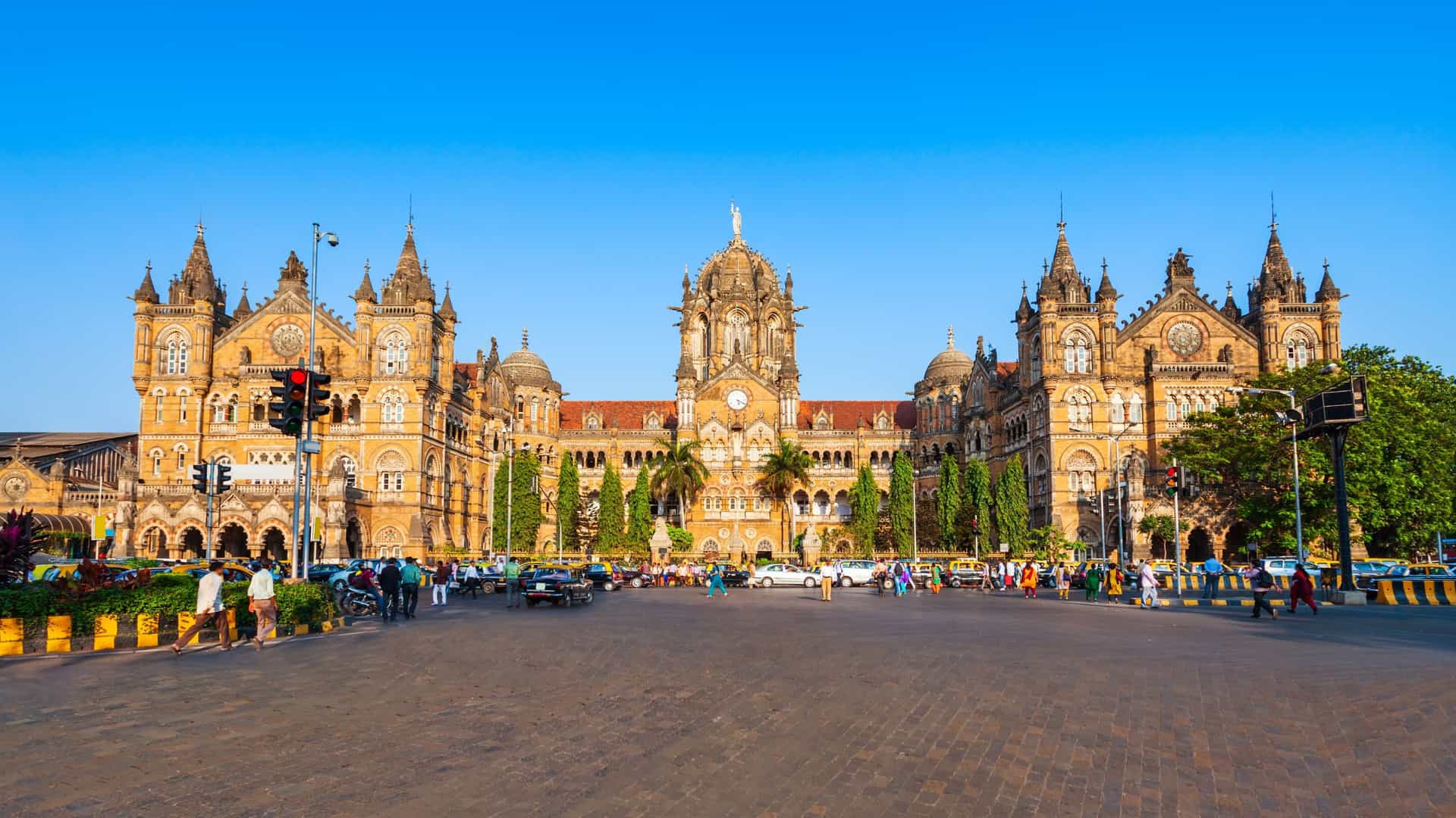  A view of the Chhatrapati Shivaji Maharaj Terminus, a UNESCO World Heritage Site in Mumbai, India. The image captures the historic Victorian Gothic Revival and traditional Indian architectural style of the train station's facade, including its intricate carvings, a large central dome topped with a statue, and multiple spires and turrets. The building is made of yellow and white stone, with a blue sky in the background, and street level traffic and pedestrians are visible in the foreground.
