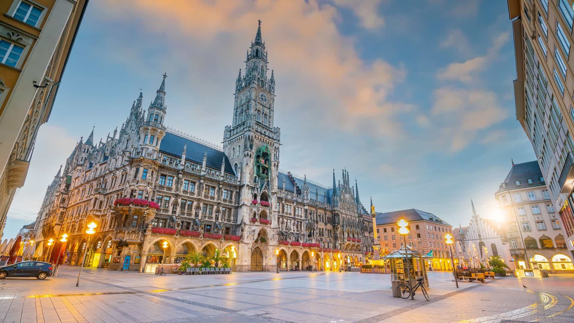  A high angle, panoramic view of the Munich downtown skyline at dusk. The city is centered on Marienplatz, with the prominent neo Gothic facade and tower of the Neues Rathaus (New Town Hall) standing out. The lights of the buildings and streets are beginning to glow, and the sky shows the soft colors of twilight.
