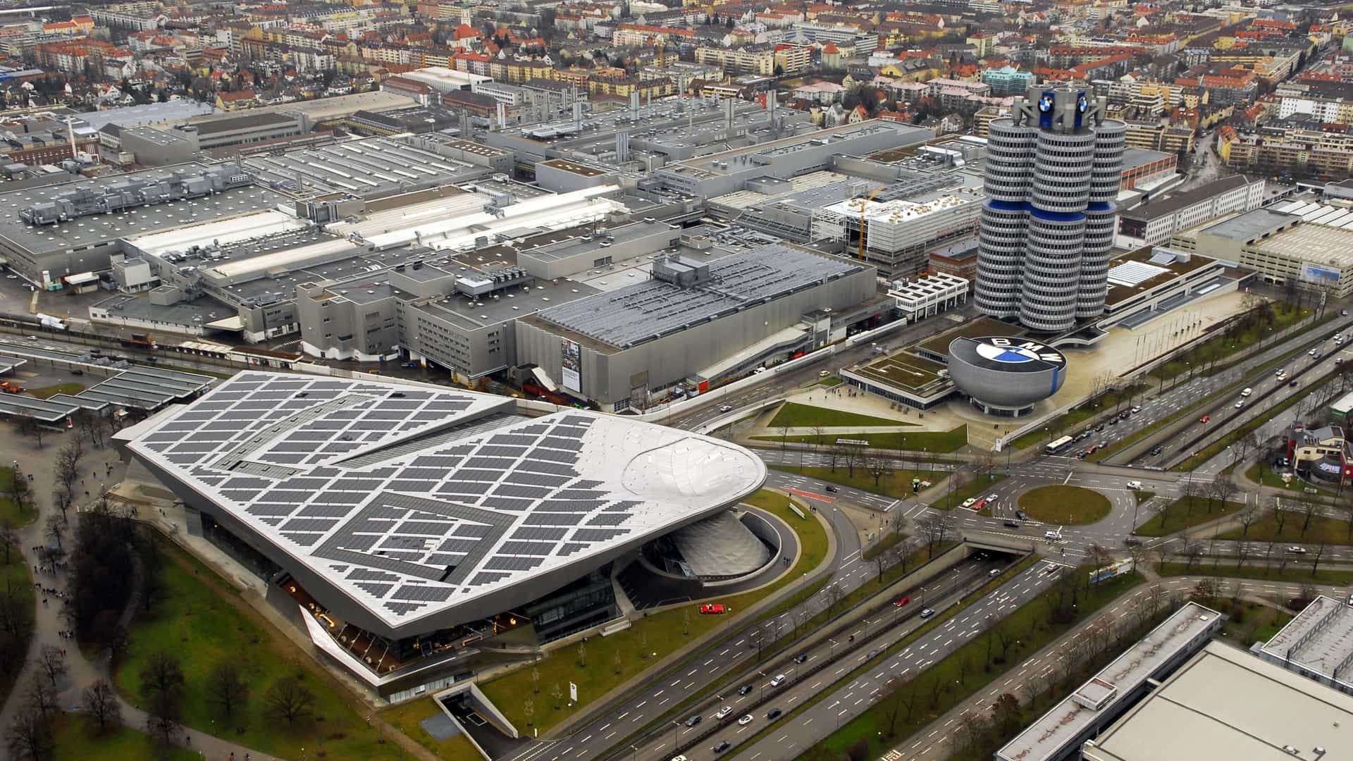 A sweeping aerial view of the modern architecture in Munich's Olympic Park district, prominently featuring the futuristic double cone design of the BMW Welt exhibition hall and the slender, towering Olympiaturm observation tower rising above the complex.