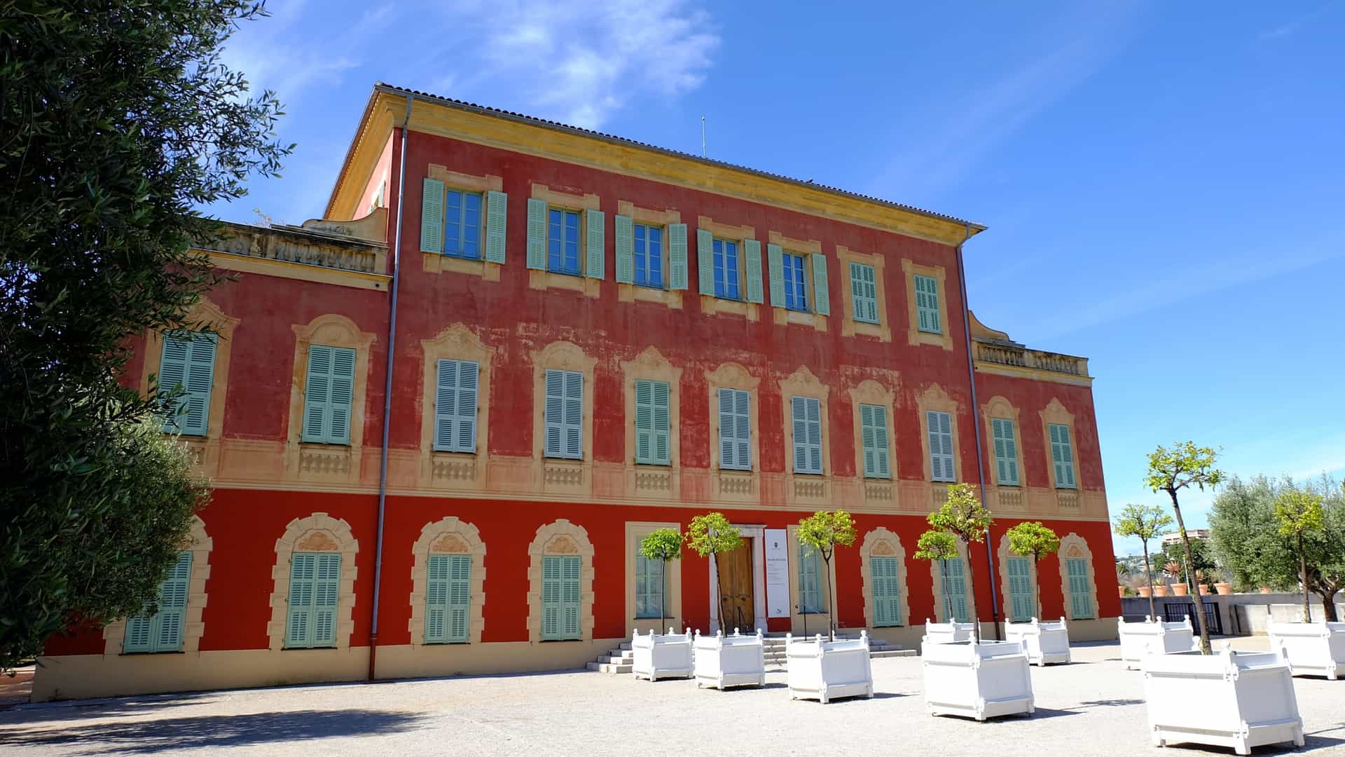 A view of the Matisse Museum (Musée Matisse) in Nice, France. The museum is a stately 17th-century Genoese villa, identifiable by its red-ochre facade, tile roof, and numerous arched windows. The building is situated on a hill within the Cimiez Gardens, surrounded by greenery and palm trees under a bright blue sky.