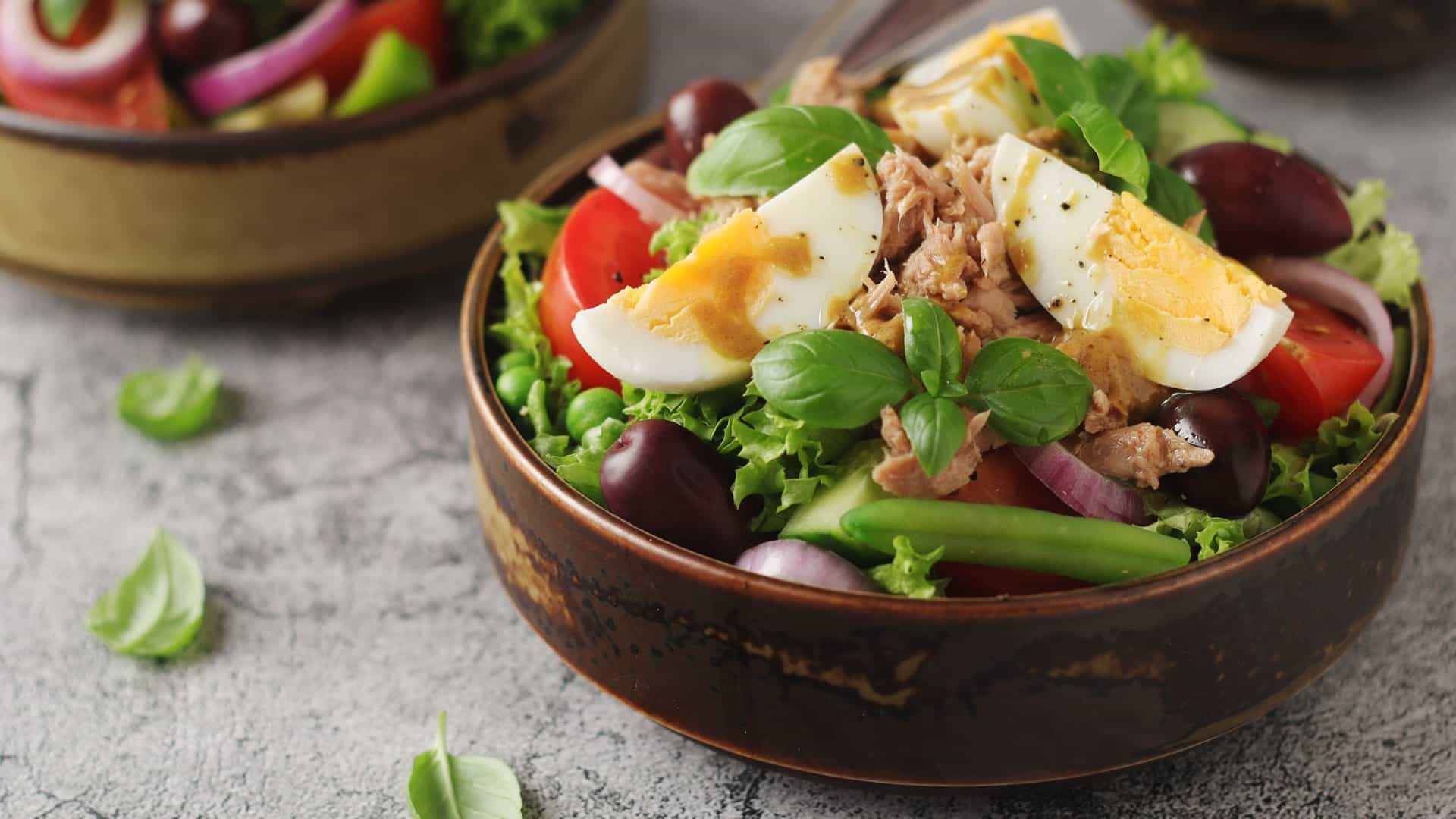 A photograph of a beautifully arranged Niçoise Salad (Salade Niçoise), a traditional French dish from Nice. The large plate contains a colorful composition of fresh ingredients, typically including sliced hard-boiled eggs, tomatoes, new potatoes, green beans, black olives, capers, and anchovies or tuna, all lightly dressed and arranged in an appealing presentation.