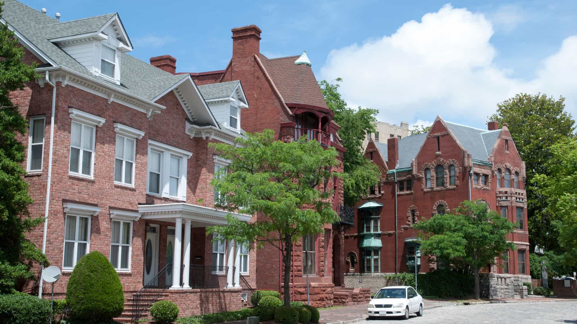 A view of the historic Freemason Street in Norfolk, Virginia, featuring a row of well-preserved, late 19th-century Victorian and Federal-style townhouses. The image highlights one or more of the houses painted a deep red or brick-red color, with white trim and architectural details like grand porches and detailed ironwork, typical of the neighborhood's elegant atmosphere.
