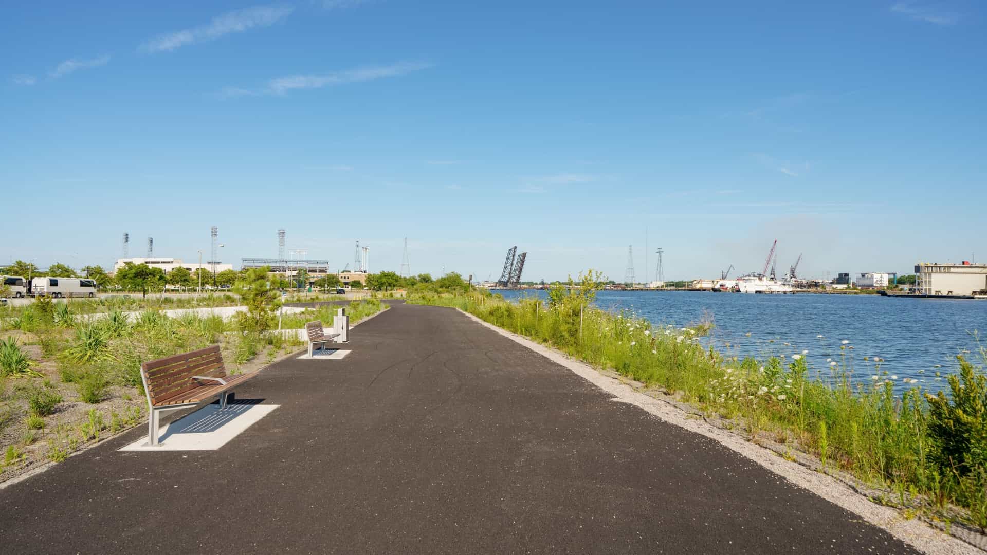 A photograph of the scenic Waterside Elizabeth Trail in Norfolk, Virginia, a paved pedestrian and cycling path running alongside the Elizabeth River. The image shows the trail lined with benches and lamp posts, offering views of the water, docked boats, and the distant Portsmouth skyline.