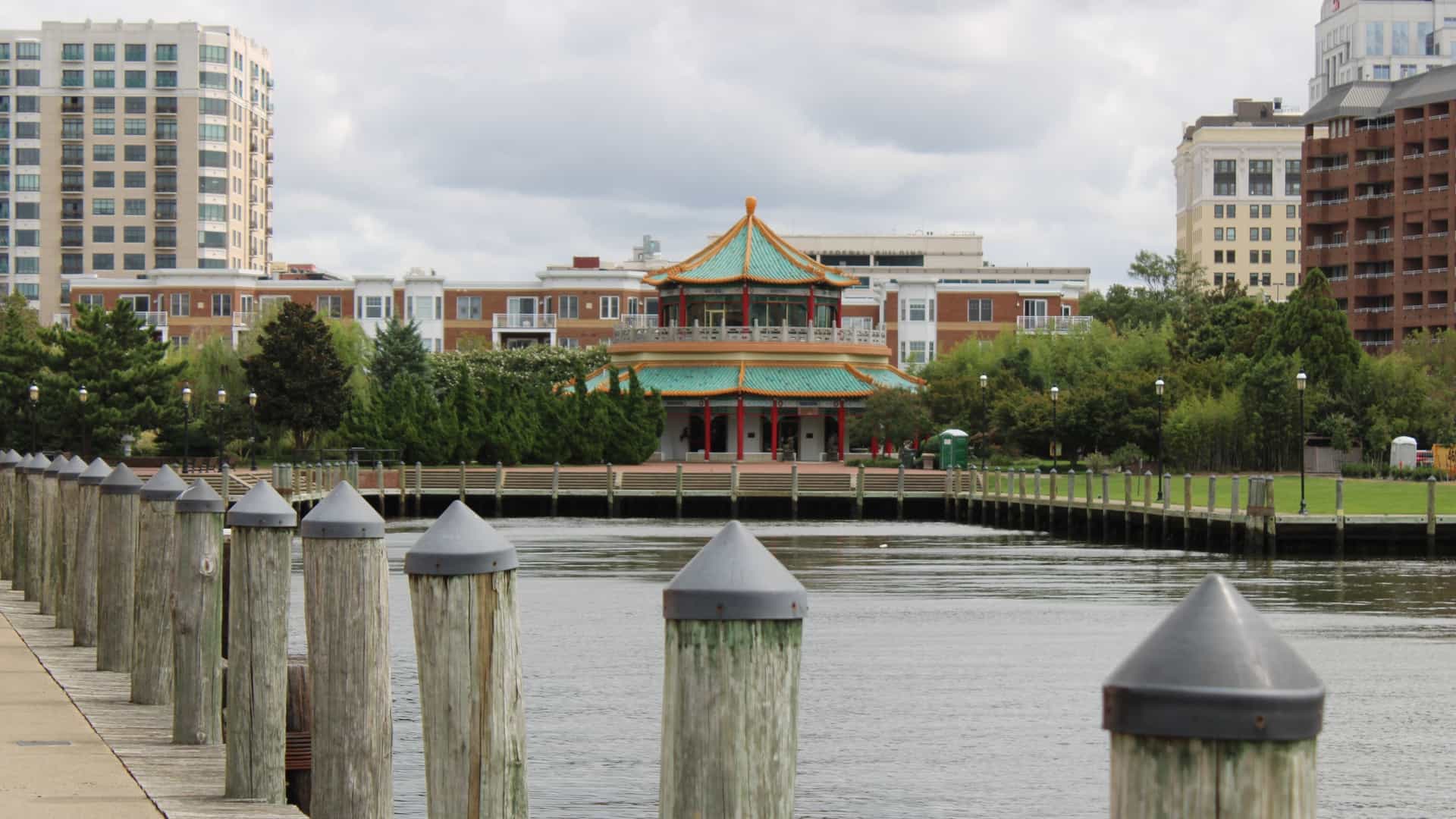 A sunny, wide-angle shot of the Downtown Norfolk Waterside District entertainment and dining complex, situated prominently along the Elizabeth River waterfront. The modern building features large windows and outdoor seating, with boats and the harbor visible in the foreground. Enjoy Local Cuisine at Waterside District.