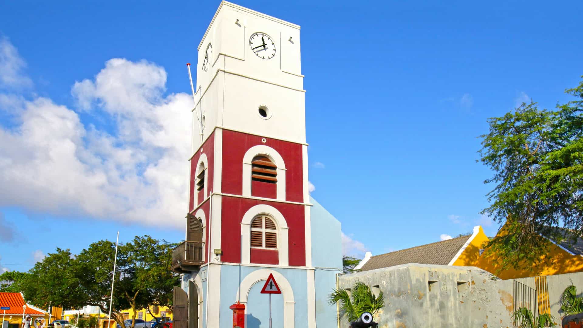 The Willem III Tower at Fort Zoutman in Oranjestad, Aruba. The historic Dutch colonial clock tower is painted in white, red, and light blue. It stands near a cobblestone street, next to a small, dark-colored cannon and a classic red post box, under a bright blue sky with white clouds. This building is the oldest structure on the island and now houses the Historical Museum of Aruba.