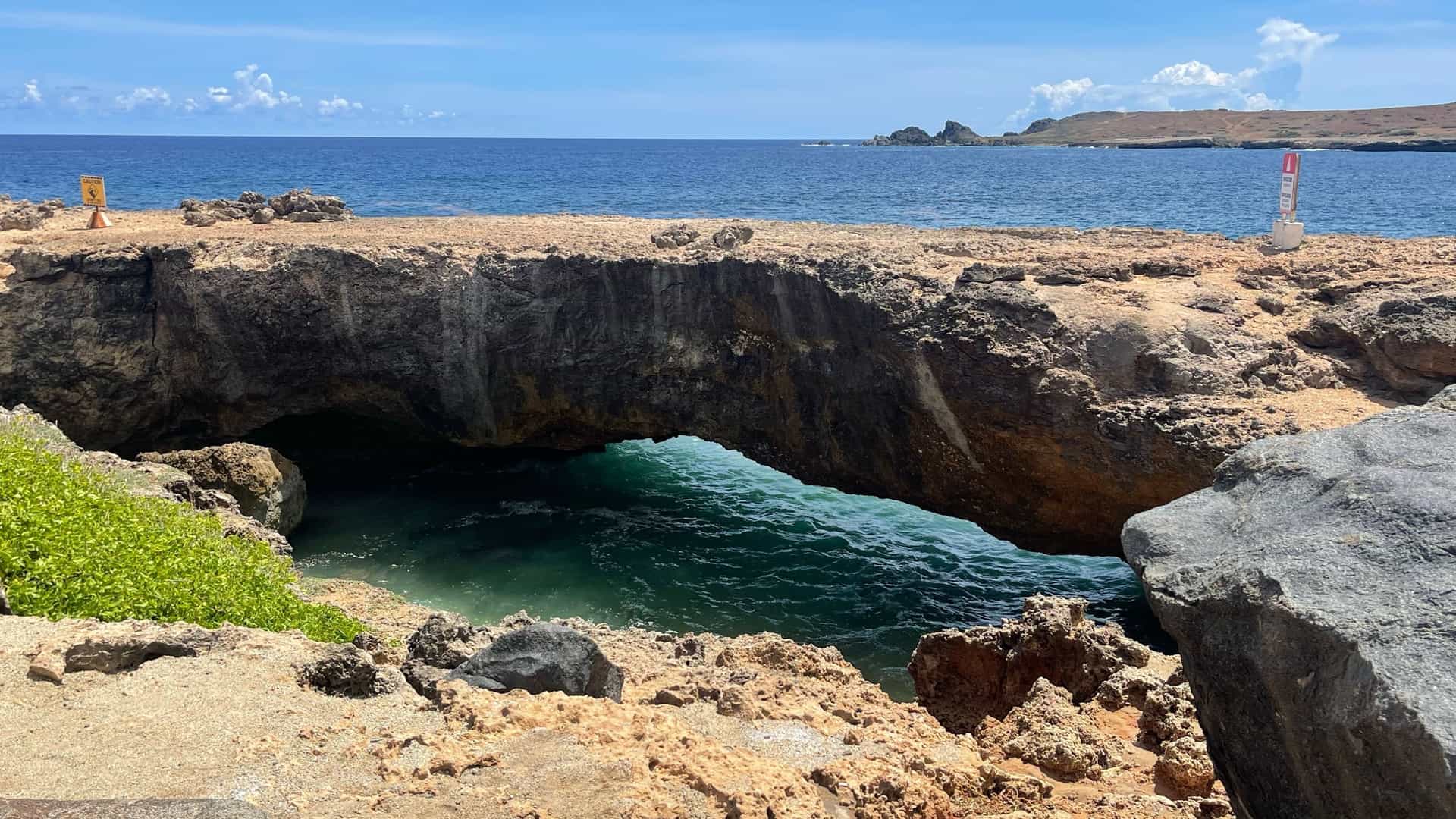 A picturesque view of the "Baby Natural Bridge" on the rugged coastline of Aruba. The smaller natural arch, formed from coral rock, stands over the turquoise Caribbean Sea. Waves crash against the black, rocky shore, under a clear blue sky, capturing the dramatic scenery of the island's eastern side in Arikok Natural Park.