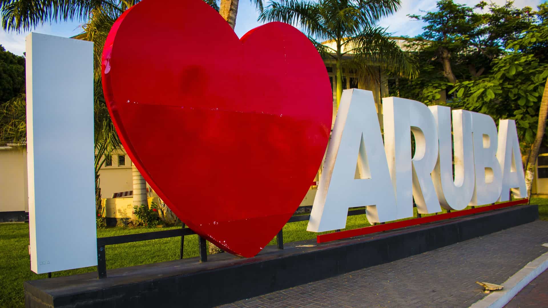 The famous "I ❤️ Aruba" sign, located in the capital city of Oranjestad, Aruba. The sculpture features large, white capital letters spelling out "I" and "ARUBA," with a large, glossy red heart replacing the word "Love." The sign stands on a stone or paved surface, with palm trees and colorful buildings in the background, promoting the island's tourism slogan, "One Happy Island."