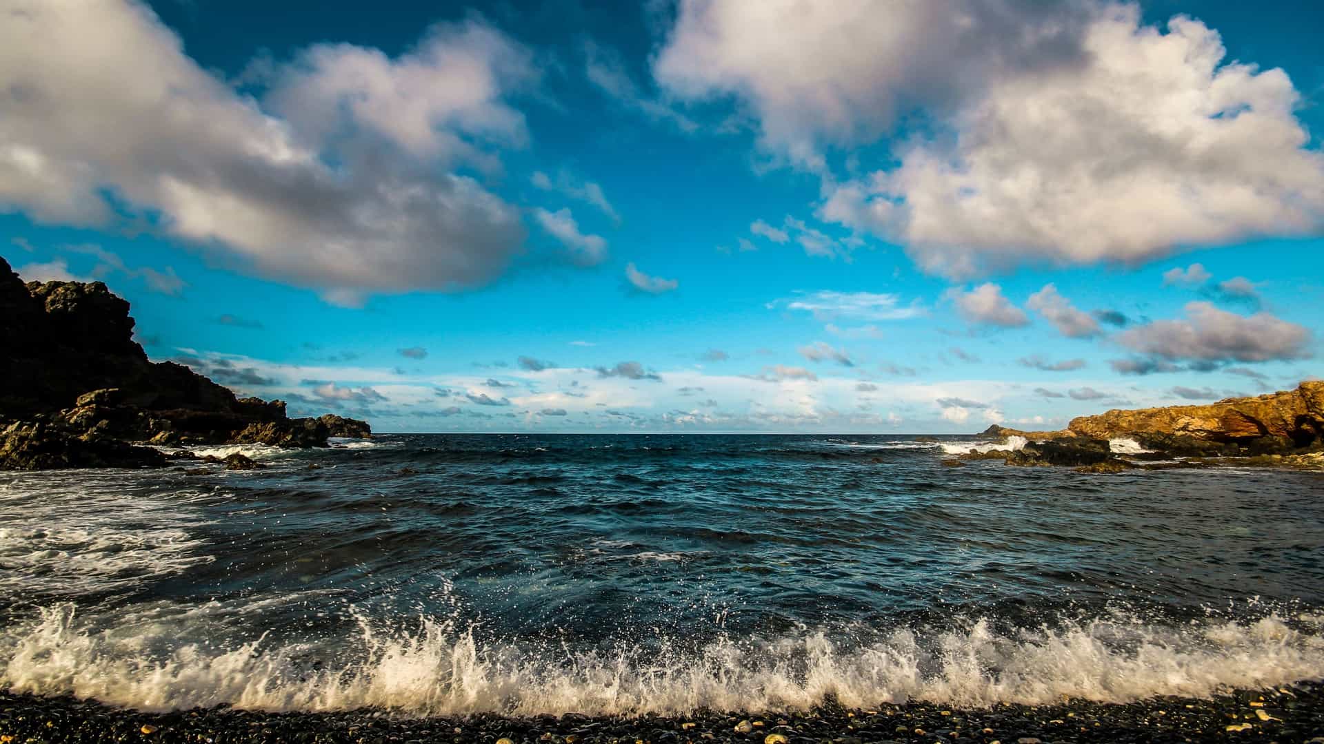 The rugged landscape of Blackstone Beach on the northeast coast of Aruba. The foreground is covered in smooth, black volcanic or coral stones, giving the beach its name. Waves from the deep blue Caribbean Sea crash against the shore, and in the distance, a small, natural rock arch or bridge is visible, with a few low, scrubby desert plants dotting the sandy background.