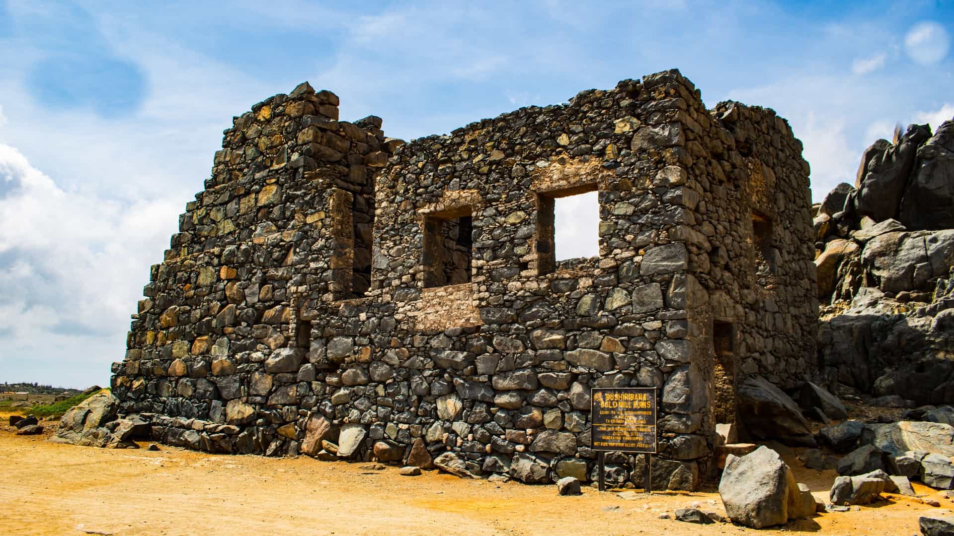 The ruins of the Bushiribana Gold Smelter on the rugged, arid northern coast of Aruba. The large, stone walls of the 19th-century gold mill stand out against the blue Caribbean Sea and a rocky, desert landscape. The remnants of the structure, featuring arched windows and doorways, are a popular tourist landmark in the Arikok National Park area.