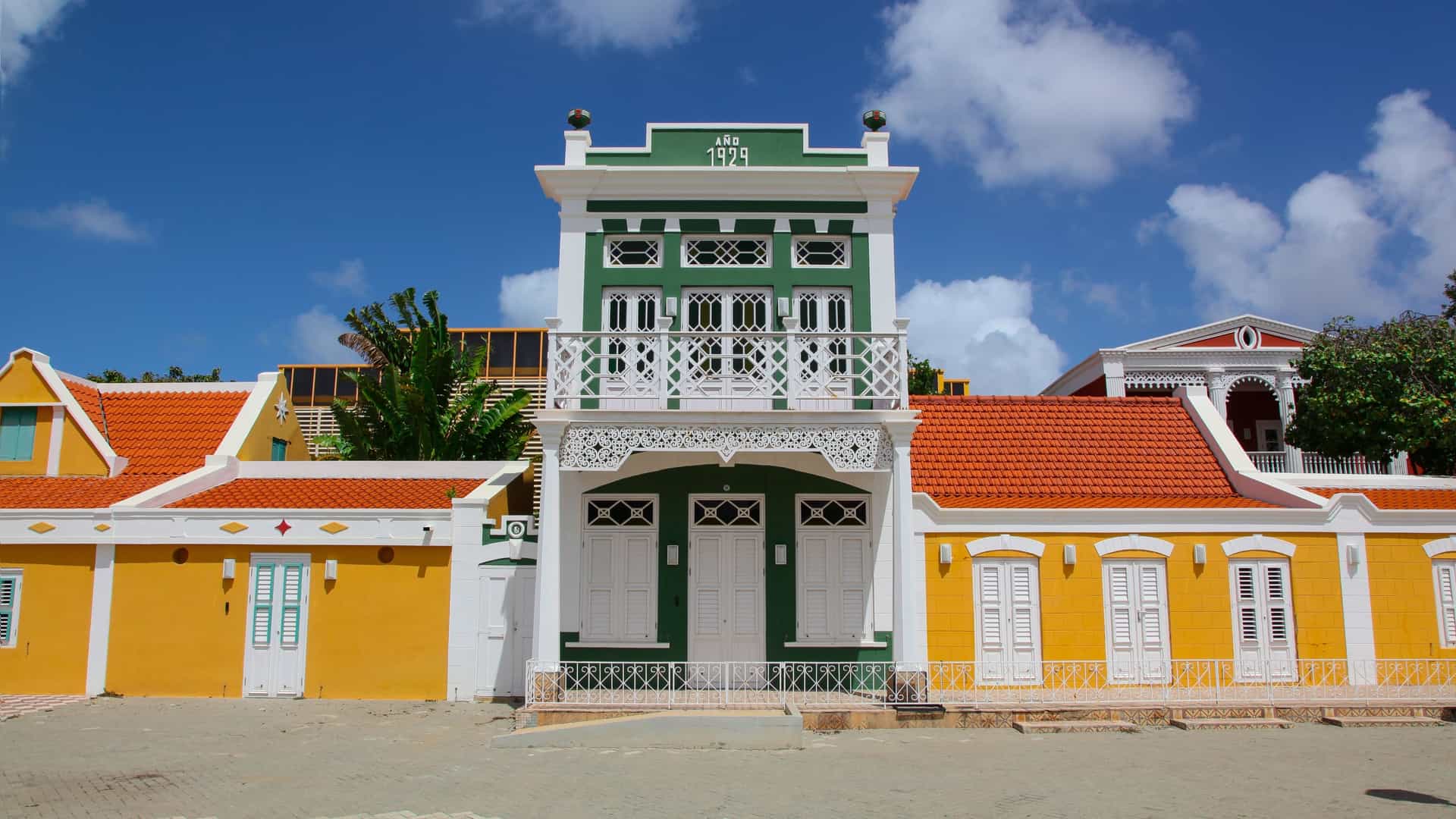 The colorful facade of the National Archeological Museum Aruba (NAMA) in Oranjestad. The historic, two-story colonial building is painted in striking colors, featuring a yellow base, a light green upper floor, and white trim around the arched windows. The entrance is topped with an elaborate, stepped Dutch-style gable, typical of the historic architecture in Aruba's capital.