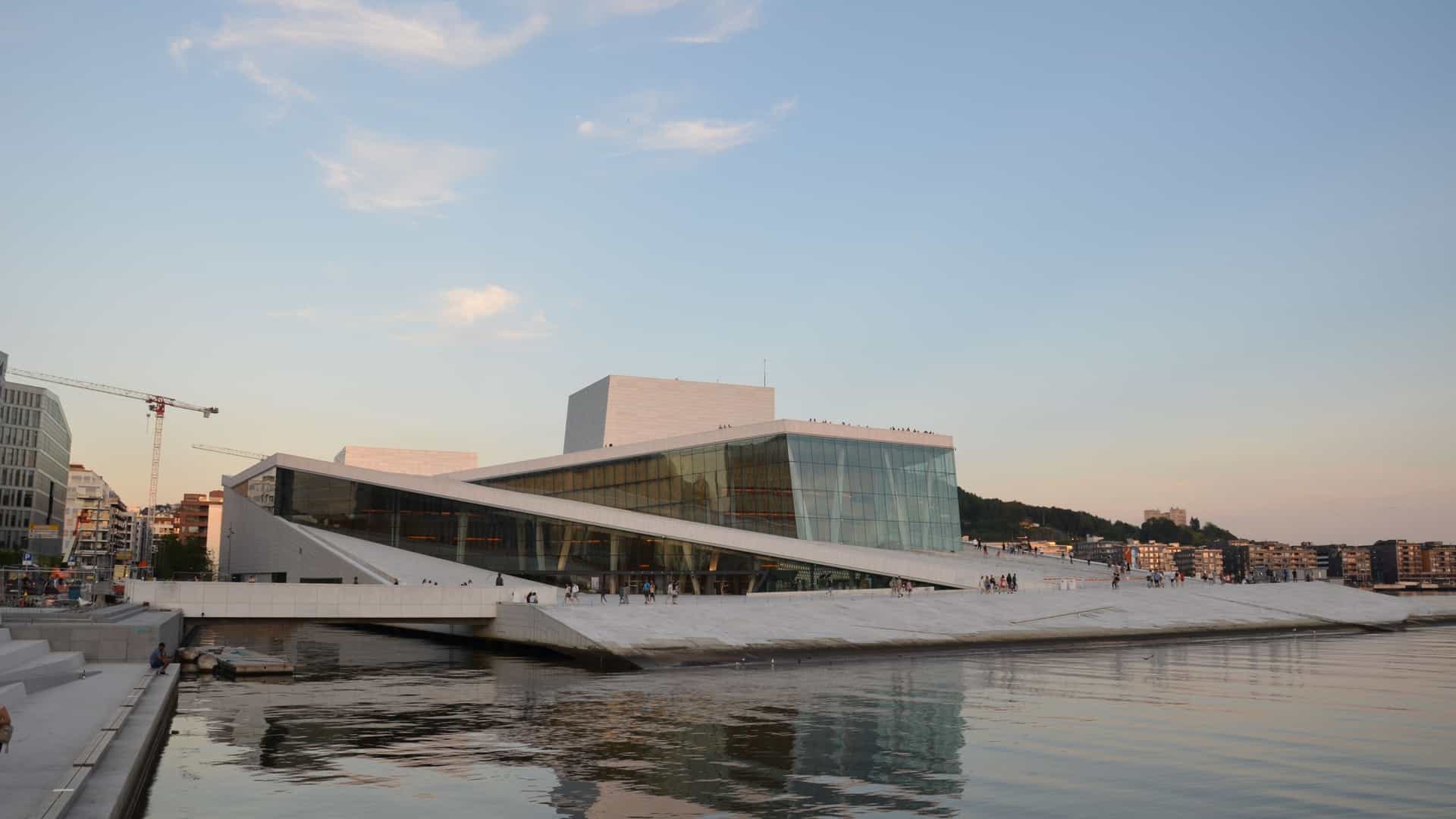  The Oslo Opera House, a modern building designed by Snøhetta, is shown from an exterior perspective. The building's defining feature is its sloping, white roof, which acts as a public plaza, appearing to rise from the waters of the Oslofjord. The exterior is clad in white Italian Carrara marble and granite. Large glass facades are visible, reflecting the sky and the surrounding cityscape. The architecture is a prominent landmark, inviting visitors to walk on its surface and seamlessly connecting the city to the fjord.