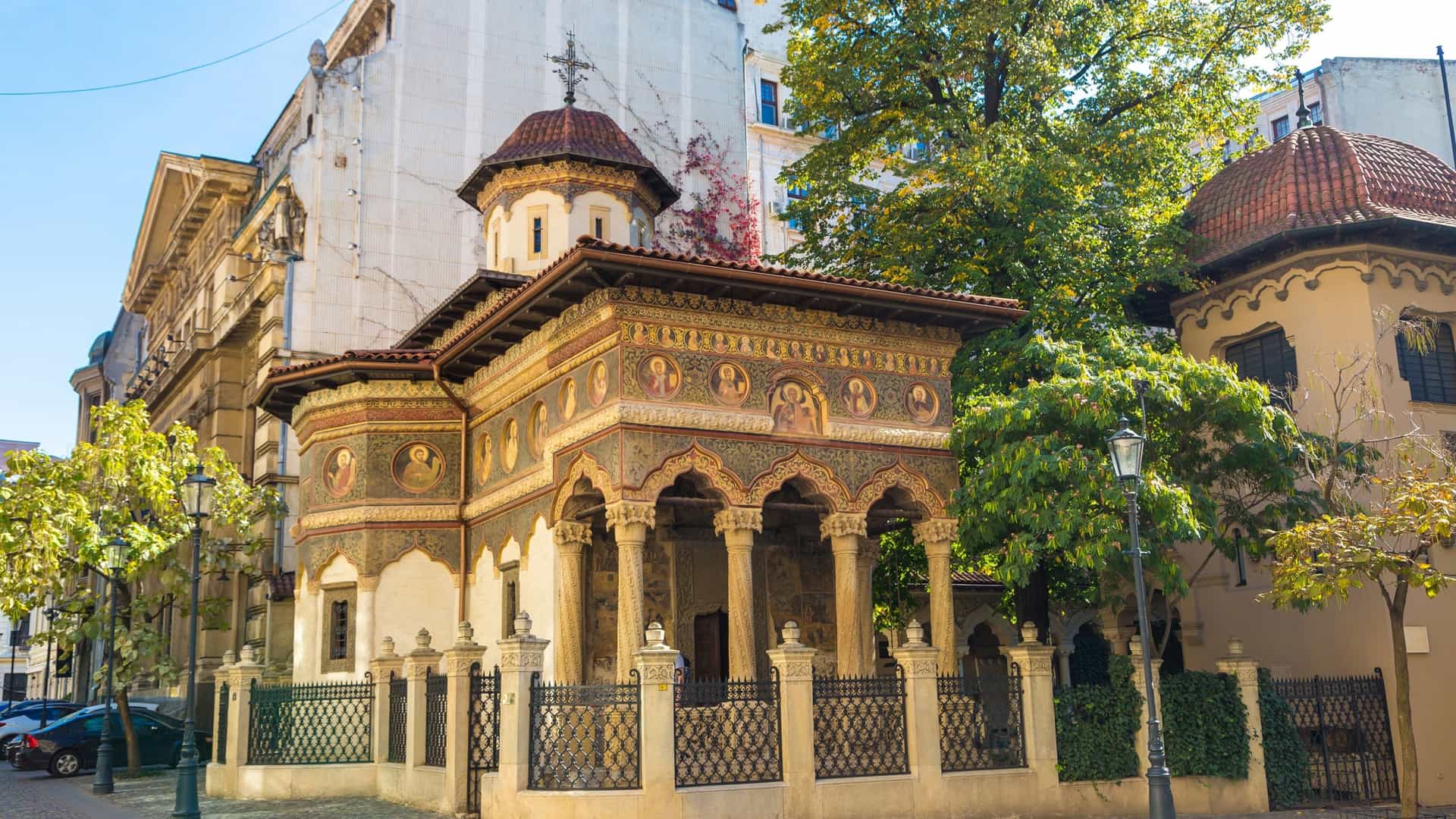 The ornately painted interior of the Stavropoleos Monastery Church (Saints Michael and Gabriel) in Bucharest, Romania. The view shows the main nave, where elaborate Byzantine frescoes cover the walls and arched ceiling, illuminated by natural light filtering through small windows.