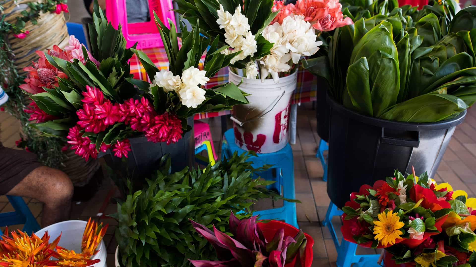 A vibrant collection of fresh tropical flowers and garlands for sale at a stall in the Papeete Market (Le Marché de Papeete) in Tahiti, French Polynesia. The stall is overflowing with colorful, fragrant bouquets and leis (flower necklaces/crowns) made from a variety of Polynesian flowers, such as the white Tiare Tahiti (Tahitian gardenia), yellow and red hibiscus, and Plumeria. The flowers are brightly lit, showcasing the abundance and central role of fresh flowers in Tahitian culture and the market's lively atmosphere.