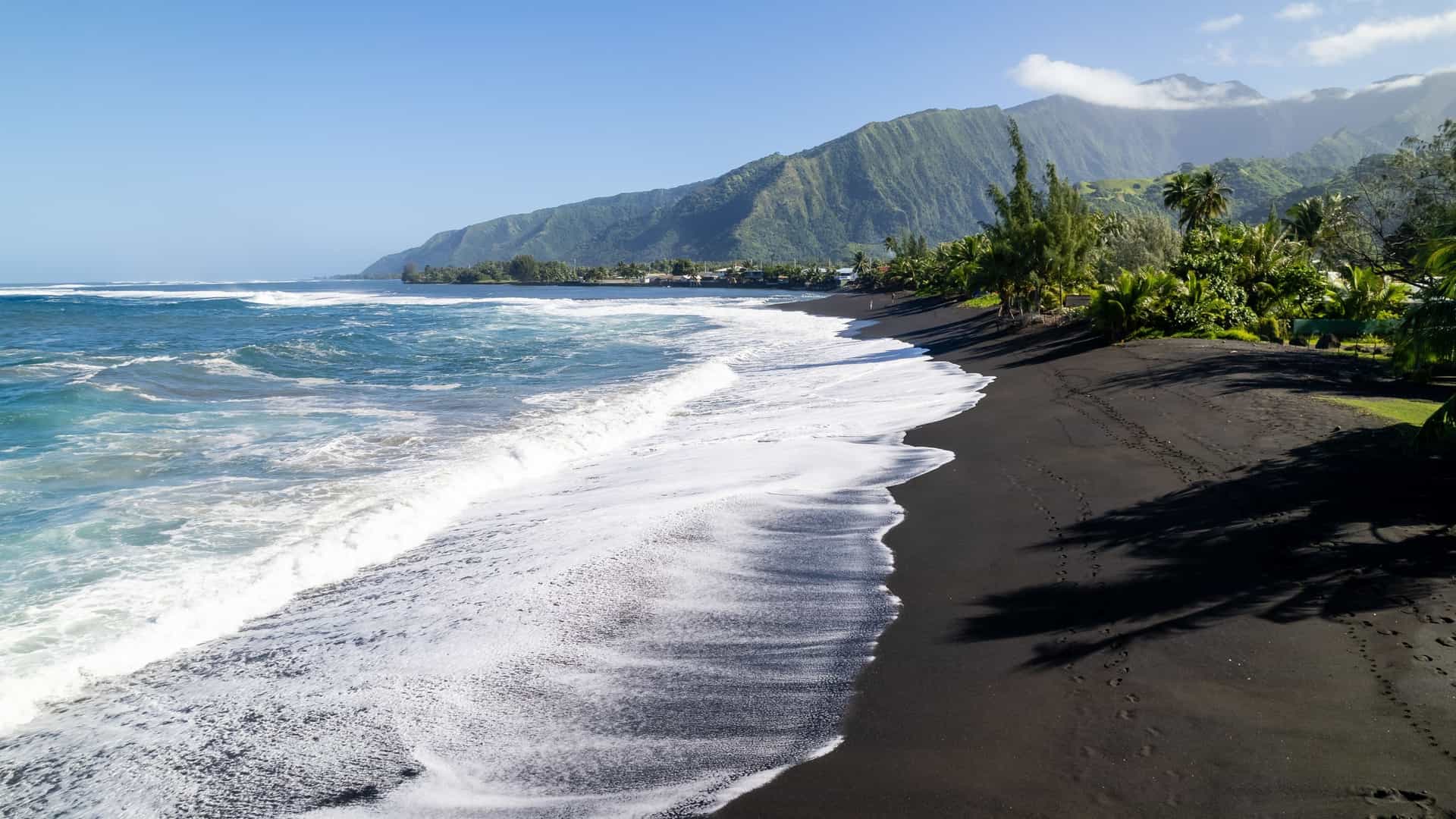 An aerial photograph of the waves of the Pacific Ocean rolling onto the black sand of Taharuu Beach in Papara, Tahiti, French Polynesia. The image captures the contrast between the white foam of the waves, the dark volcanic sand of the beach, and the lush green vegetation and volcanic mountains visible in the background.