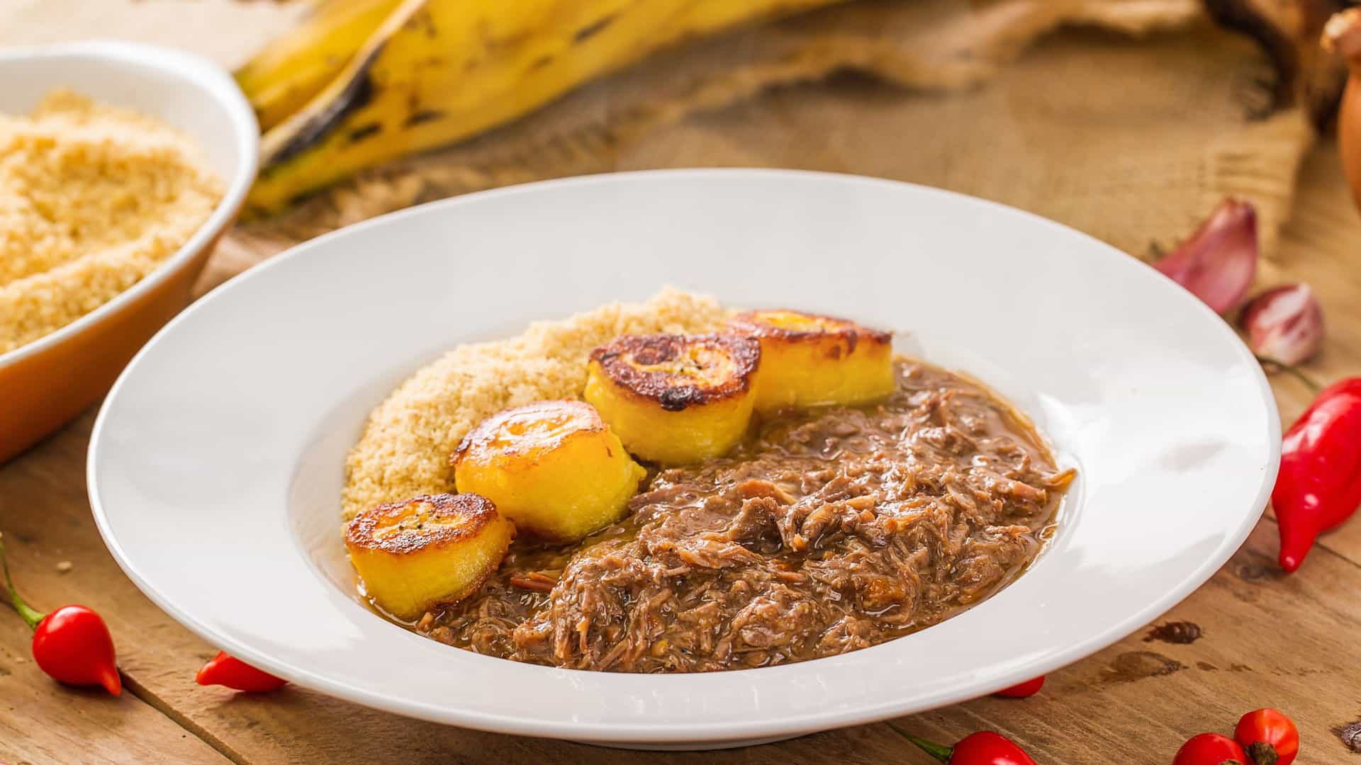 A plate of Barreado, a traditional Southern Brazilian meat stew from Paraná. The dish consists of finely shredded, long-cooked beef served alongside a generous mound of dry manioc flour (farinha de mandioca), which is used to thicken the stew's savory broth into a paste. The plate is also garnished with slices of banana, a classic accompaniment that adds a touch of sweetness. The stew is known for its cooking method where it's sealed in a clay pot and slow-cooked for many hours.