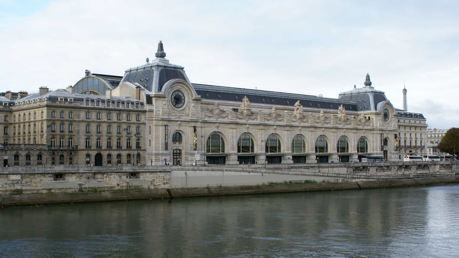 Panoramic view of the Musée d'Orsay on the Left Bank of the River Seine in Paris. The building, a former Beaux-Arts railway station, is distinguishable by its classical stone façade, monumental arched entrance, and glass roof. The river's bank is lined with trees and stone embankments, with the museum standing prominently on the left side of the frame as seen from across the water.
