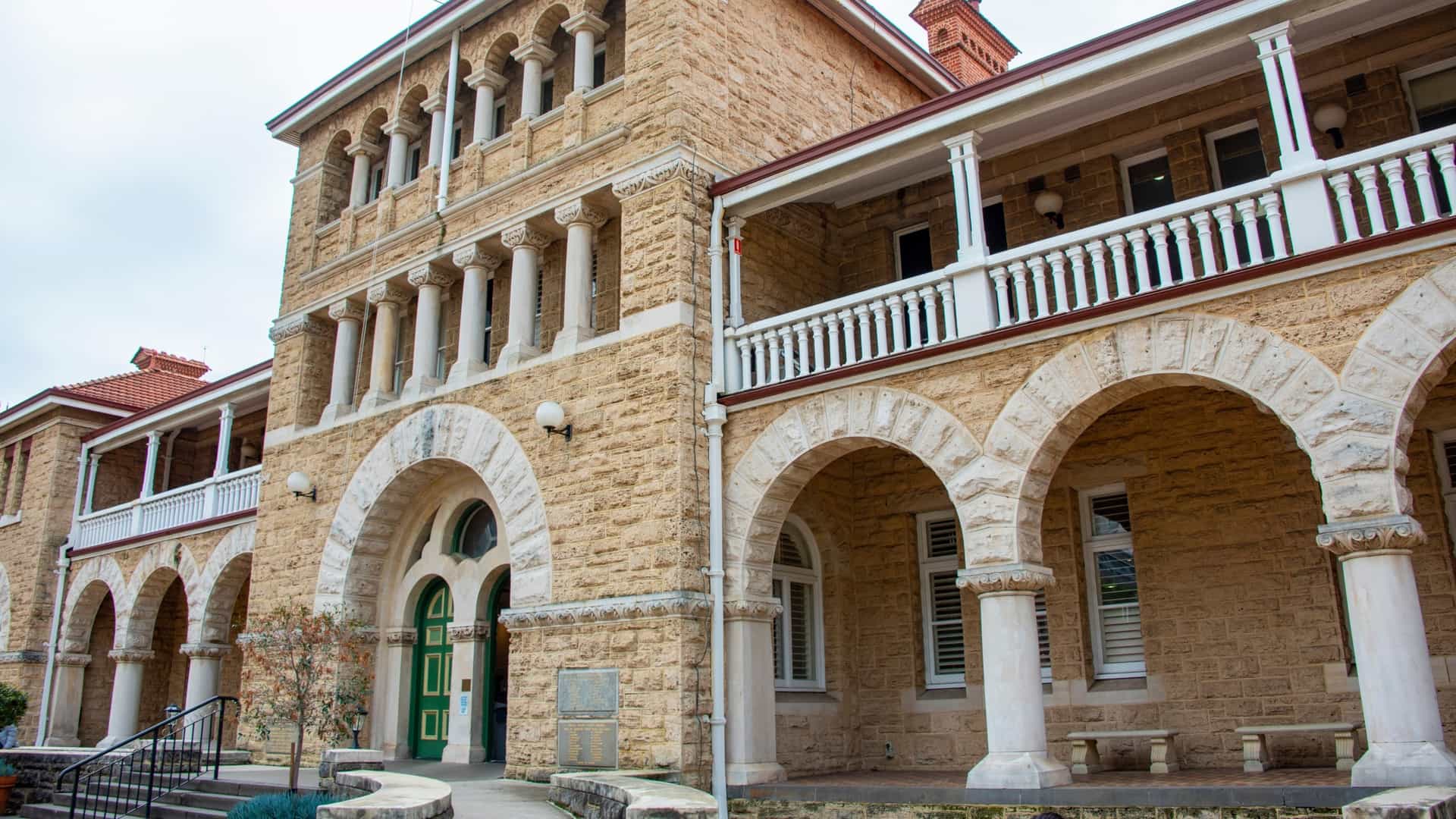 A clear photograph of the historic Perth Mint building, a stately two-story structure featuring a classical Federation Free Classical architectural style. The building is made of golden-yellow stone, with a prominent central entrance and tall windows, set against a backdrop of modern high-rise buildings and blue sky, highlighting the contrast between Perth's history and its contemporary downtown.