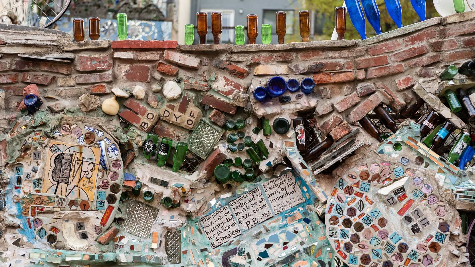 A vibrant and colorful view of the Philadelphia's Magic Gardens outdoor sculpture garden. The walls, steps, and structures are completely covered in intricate mosaics made from mixed media, including colorful tile shards, glass bottles, pieces of mirror, and found objects. The artwork, which features abstract patterns and folk art imagery, creates a swirling, immersive labyrinth under the sunlight.
