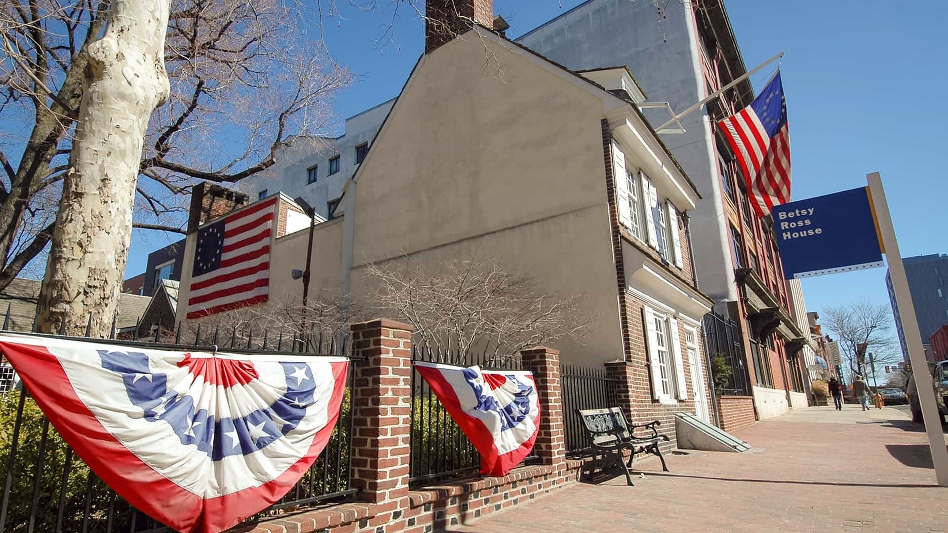 The exterior of the Betsy Ross House, a small, two-and-a-half-story historic brick row house in Philadelphia. The red brick facade features white trim around the windows and a dark green painted door. An American flag with thirteen stars (the Betsy Ross flag design) flies prominently from a pole over the entrance, marking the home of the woman credited with sewing the first American flag. The building is situated on a cobblestone street.