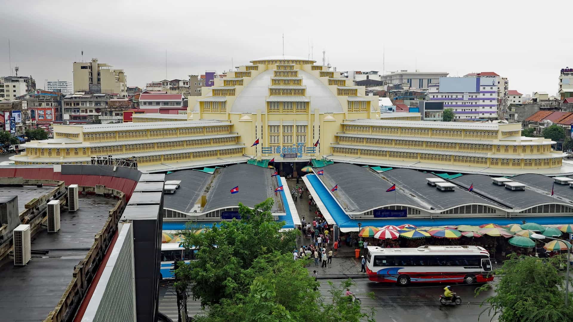 The distinct yellow-domed roof and main entrance of the Phnom Penh Central Market (Phsar Thmey) in Cambodia, seen on a rainy day. The large, Art Deco-style building is partially obscured by rain and wet pavement. Vendors' awnings and the general street activity are visible beneath the market's prominent, central yellow dome.