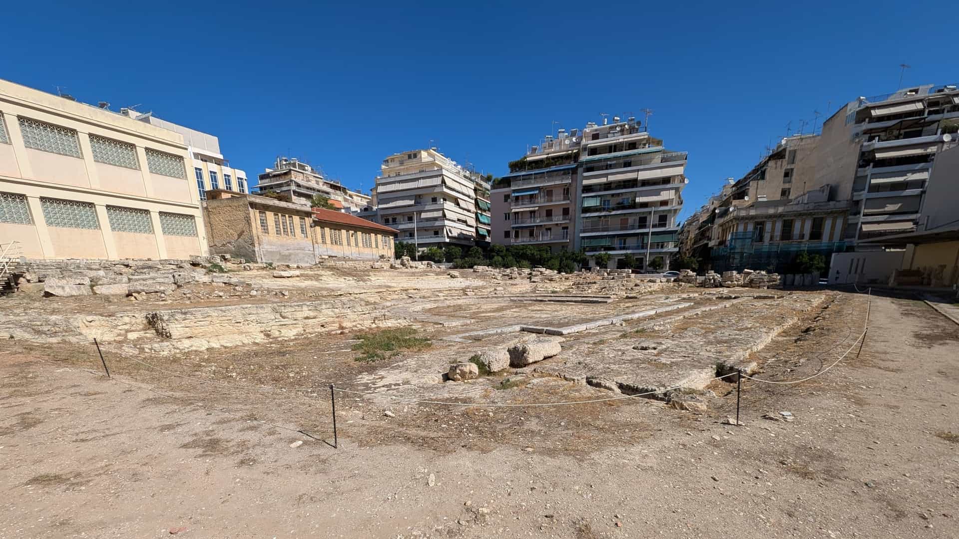 A photograph of the Archaeological Museum of Piraeus in Athens, Greece. The modern, rectangular museum building features a light-colored stone or stucco facade with large, tall windows. In the foreground, there are ancient columns and architectural fragments displayed in the outdoor courtyard, showcasing the museum's historical collection.