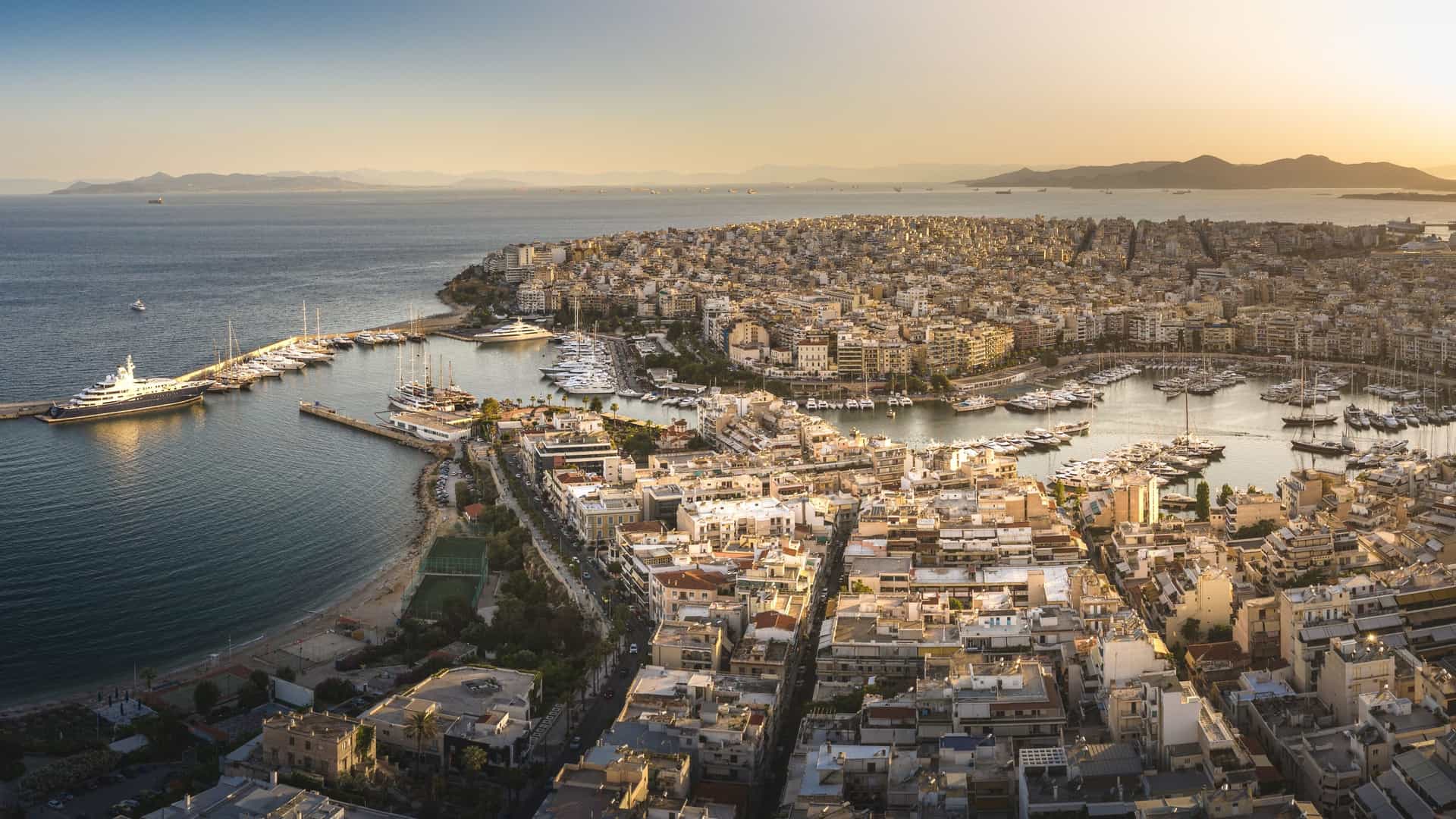 A bright, aerial drone panoramic photo of the bustling Port of Piraeus in Athens, Greece. The wide harbor is filled with numerous ferries, cruise ships, and smaller boats. The dense urban sprawl of the city of Piraeus surrounds the curved harbor, with residential and commercial buildings extending into the distance under a sunny sky.