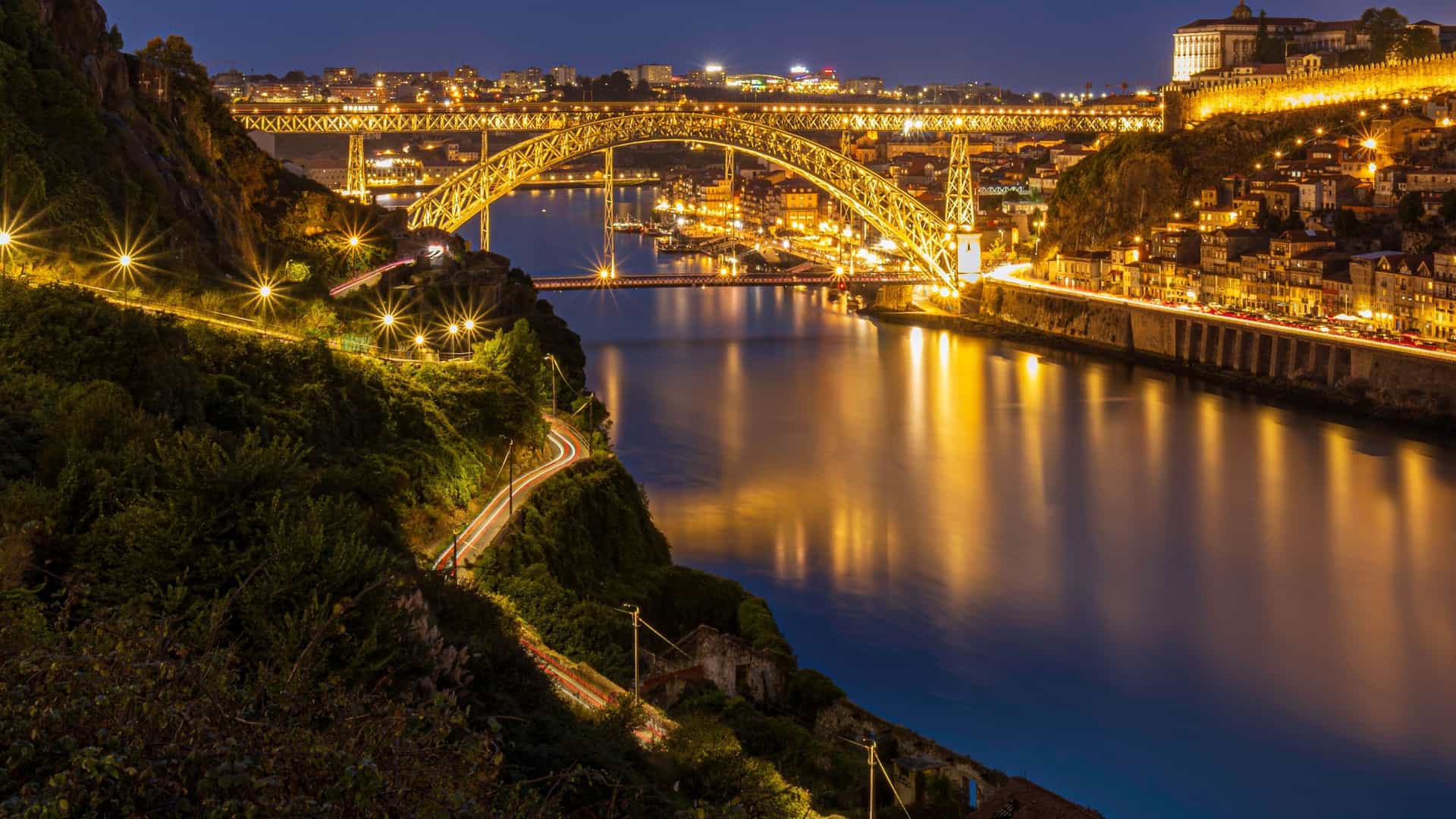 The Dom Luís I Bridge, an iconic double deck metal arch bridge, spanning the Douro River in Porto, Portugal. The wrought iron structure connects the cities of Porto and Vila Nova de Gaia, set against a bright, sunny sky.