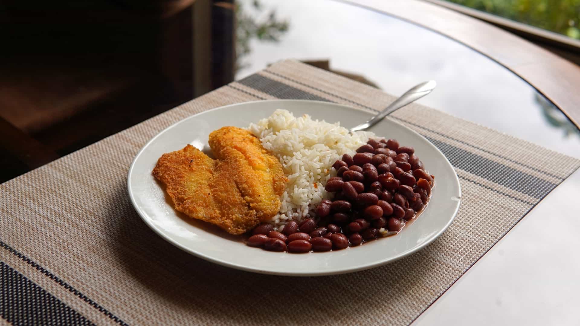  A plate of food featuring a large, golden brown breaded tilapia fillet served alongside a portion of white rice mixed with black beans (Gallo Pinto) and a side of sliced fresh tomatoes and cucumbers.