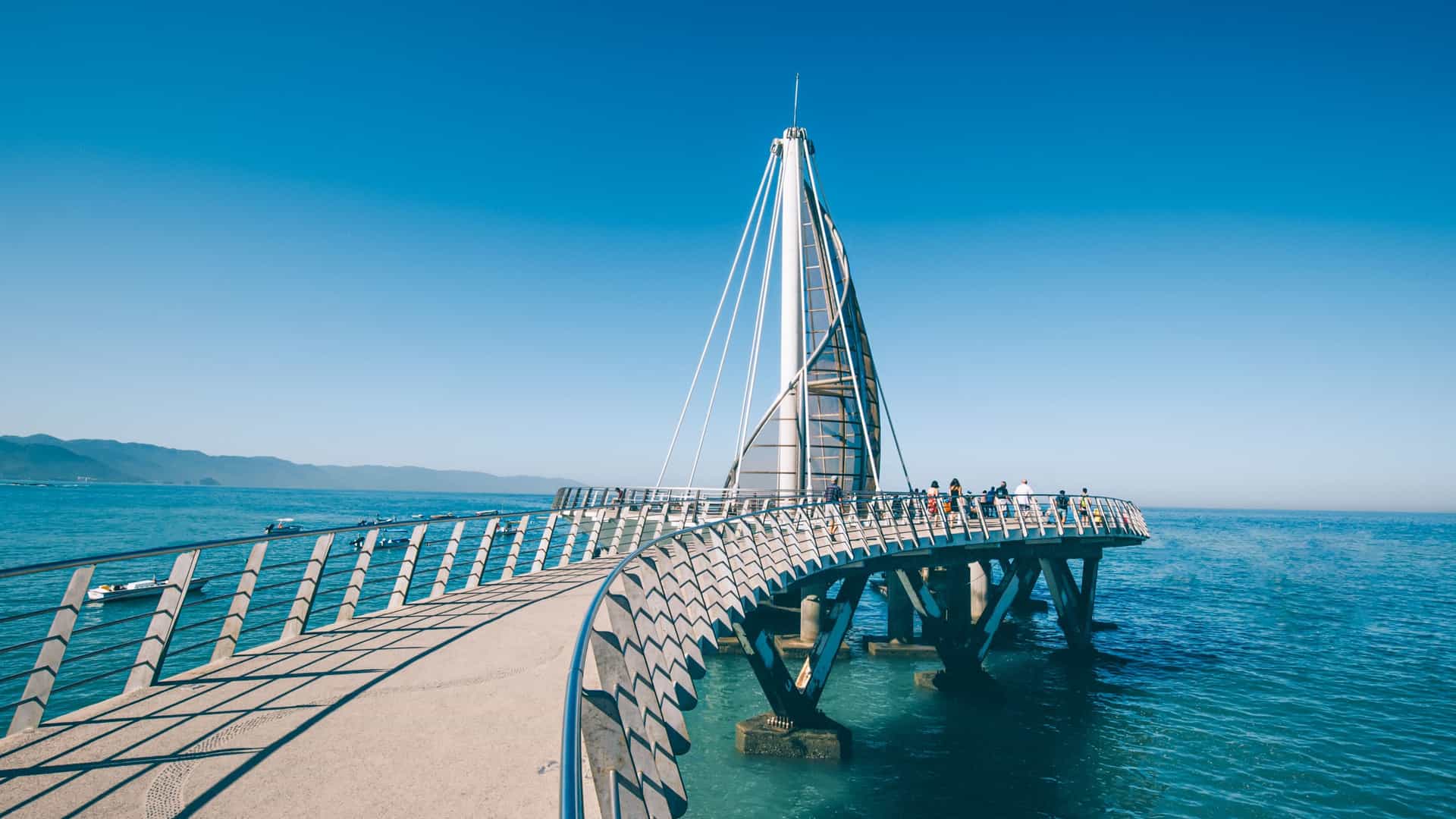 A distinctive photograph of the Muelle de Playa de los Muertos (Pier of the Beach of the Dead) in Puerto Vallarta, Mexico. The pier, a contemporary architectural structure, features a dramatically sweeping, spiral sail-like canopy at its end. The pier extends over the blue Pacific Ocean, with the sandy beach and the city's coastal buildings visible in the background under a sunny sky.
