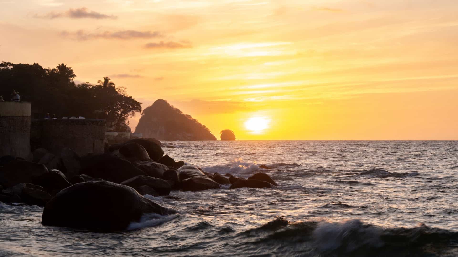 A highly dramatic photograph of the natural rock formation known as Los Arcos (The Arches), located in the Bay of Banderas near Puerto Vallarta, Mexico. The rocky islets are completely silhouetted against a brilliant sunset sky, which is filled with vibrant orange, red, and purple colors reflected on the Pacific Ocean water.