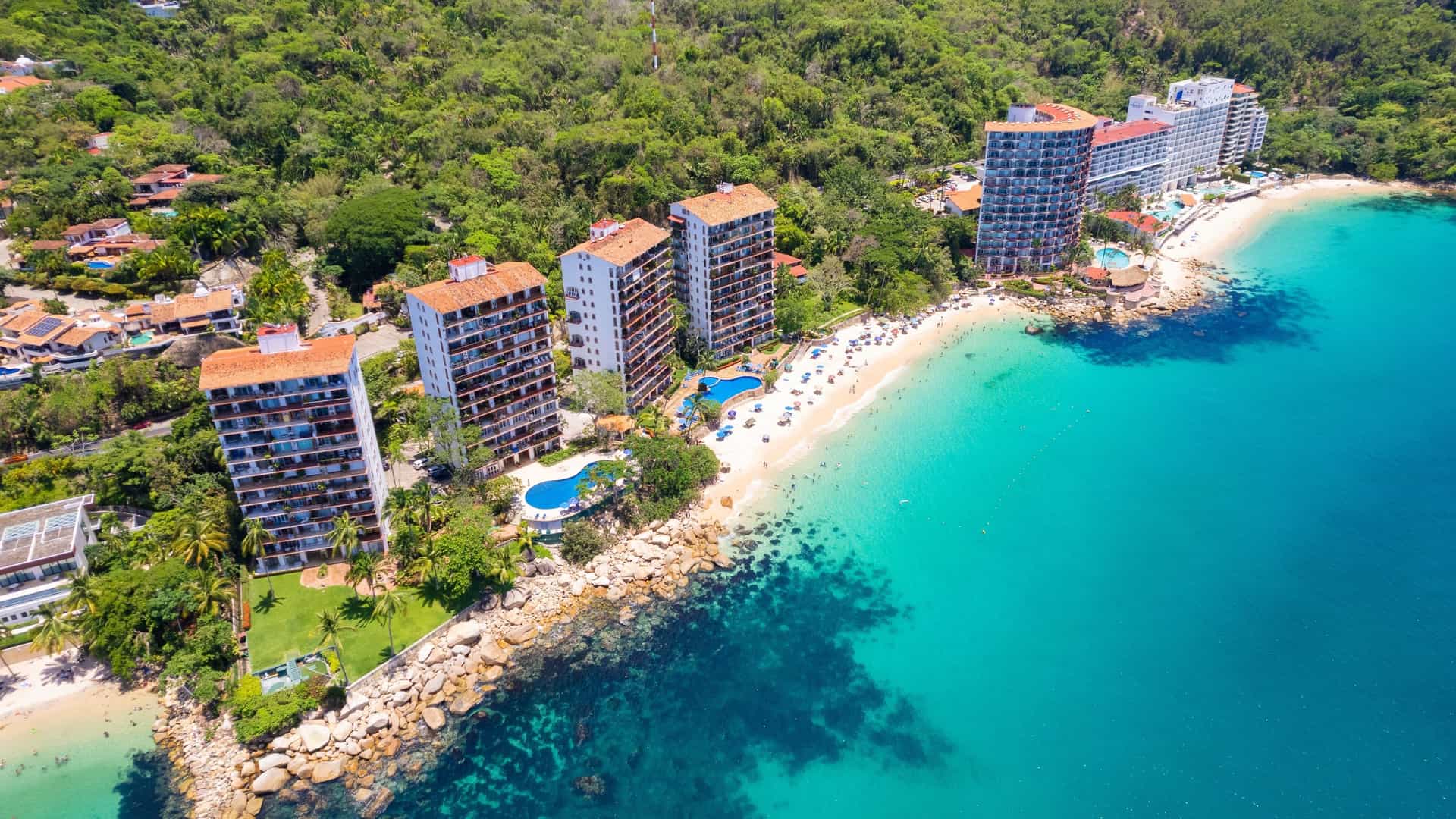 A high aerial view of the scenic Las Gemelas Beach (The Twins Beach), south of Puerto Vallarta, Mexico. The image shows two small, picturesque coves with white sand separated by a rocky outcrop. The brilliant turquoise-blue water of the Pacific Ocean contrasts sharply with the deep green, mountainous jungle terrain that rises directly behind the beaches.