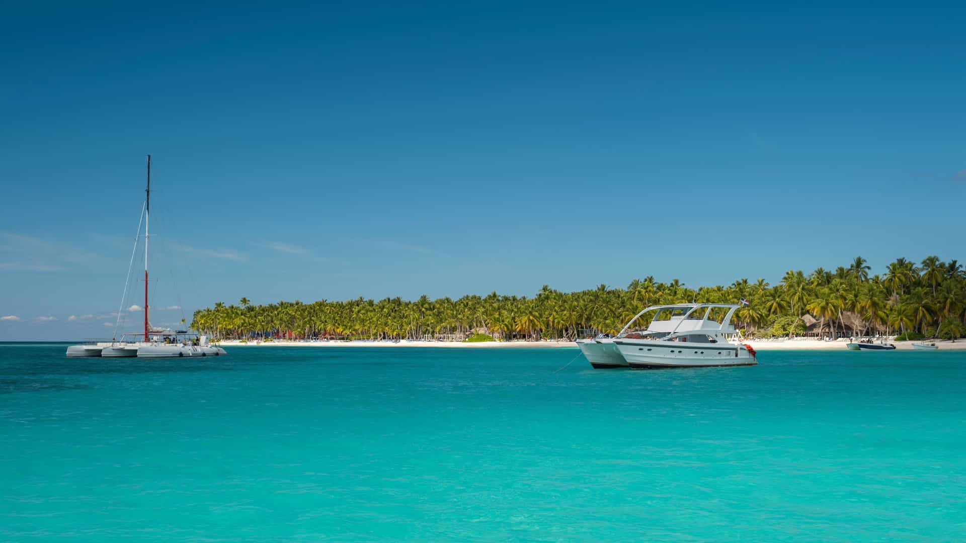 A bright, sunny photograph of a large, modern passenger catamaran sailing across dazzling turquoise-blue ocean water. The boat is full of travelers relaxing on the deck, illustrating the scenic experience of how to Take a catamaran cruise to Saona Island. The tropical sea is calm, and the large sailing vessel moves smoothly under a clear, partially cloudy sky.