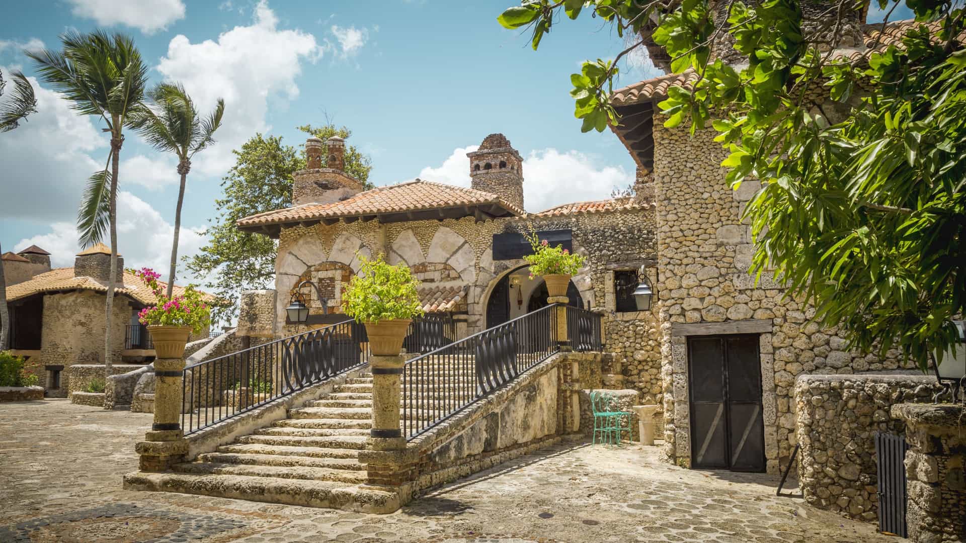 A bright, sunlit photograph showcasing the cobblestone streets and stone architecture of Altos de Chavón, a replica 16th-century Mediterranean village near La Romana, Dominican Republic. The scene features historic-looking stone buildings with tiled roofs and wrought-iron details, surrounded by palm trees and tropical landscaping, evoking the atmosphere of an old European town in a Caribbean setting.