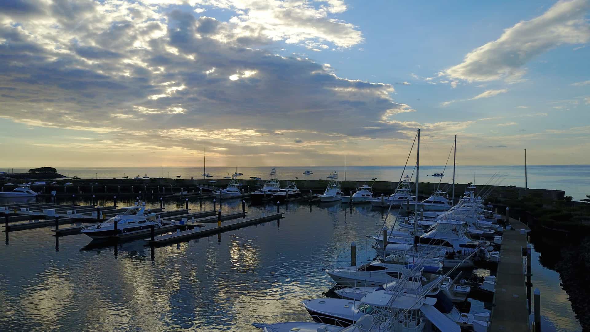 An image of Marina Pez Vela in Quepos, Costa Rica, viewed from a slightly elevated perspective. The marina basin is filled with numerous boats of various sizes, including large sportfishing yachts, catamarans, and pleasure crafts, all docked at the modern, floating concrete slips. Lush, rainforest-covered hills form a dramatic backdrop to the facility, which also features commercial buildings and waterfront restaurants along the boardwalk. The water in the harbor is a calm, dark blue-green.