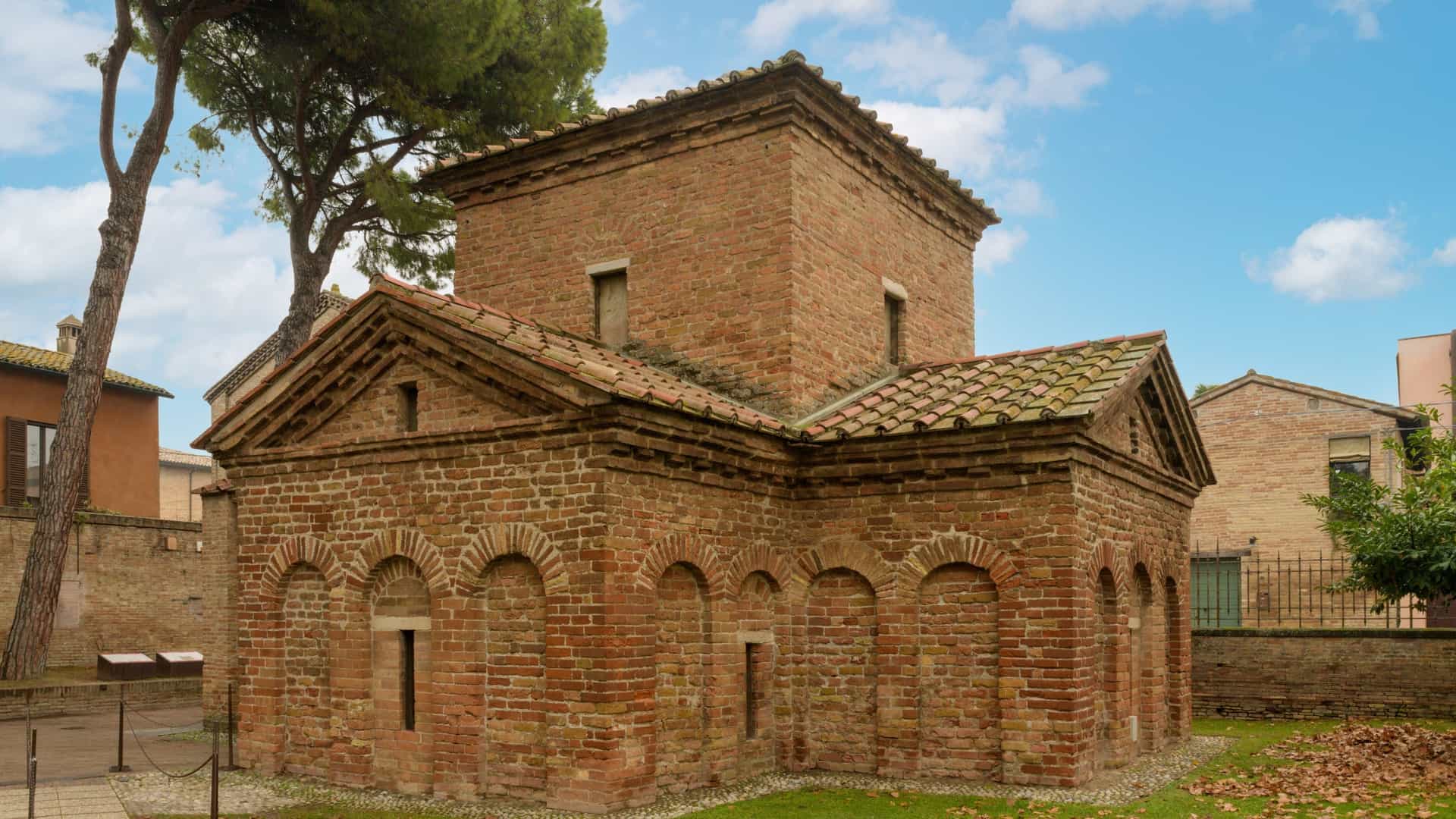  The interior of the Mausoleum of Galla Placidia in Ravenna, Italy, showing the opulent mosaics that cover the vaulted ceiling. The dome features a deep indigo blue background with a pattern of golden stars surrounding a central golden cross, with the four symbols of the Evangelists visible in the corner pendentives. The walls below are decorated with mosaics of lush vines and flowers.