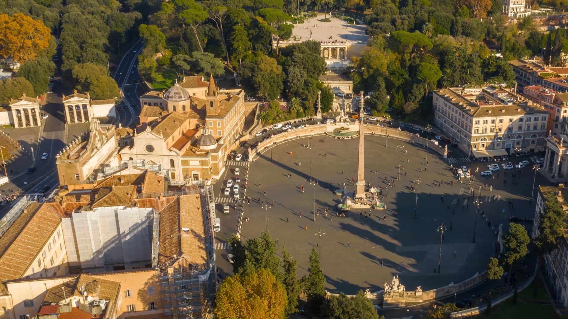  An aerial view of the Piazza del Popolo, a large, elliptical neoclassical square in Rome. The ancient Egyptian Flaminio Obelisk stands at the center, surrounded by the Fontana dei Leoni (Fountain of the Lions). At the southern end, the two nearly identical domed churches, Santa Maria in Montesanto and Santa Maria dei Miracoli, frame the start of the three streets forming the Trident. The square is bordered by historic buildings and connected to the lush, tree covered Pincio Hill on the right.