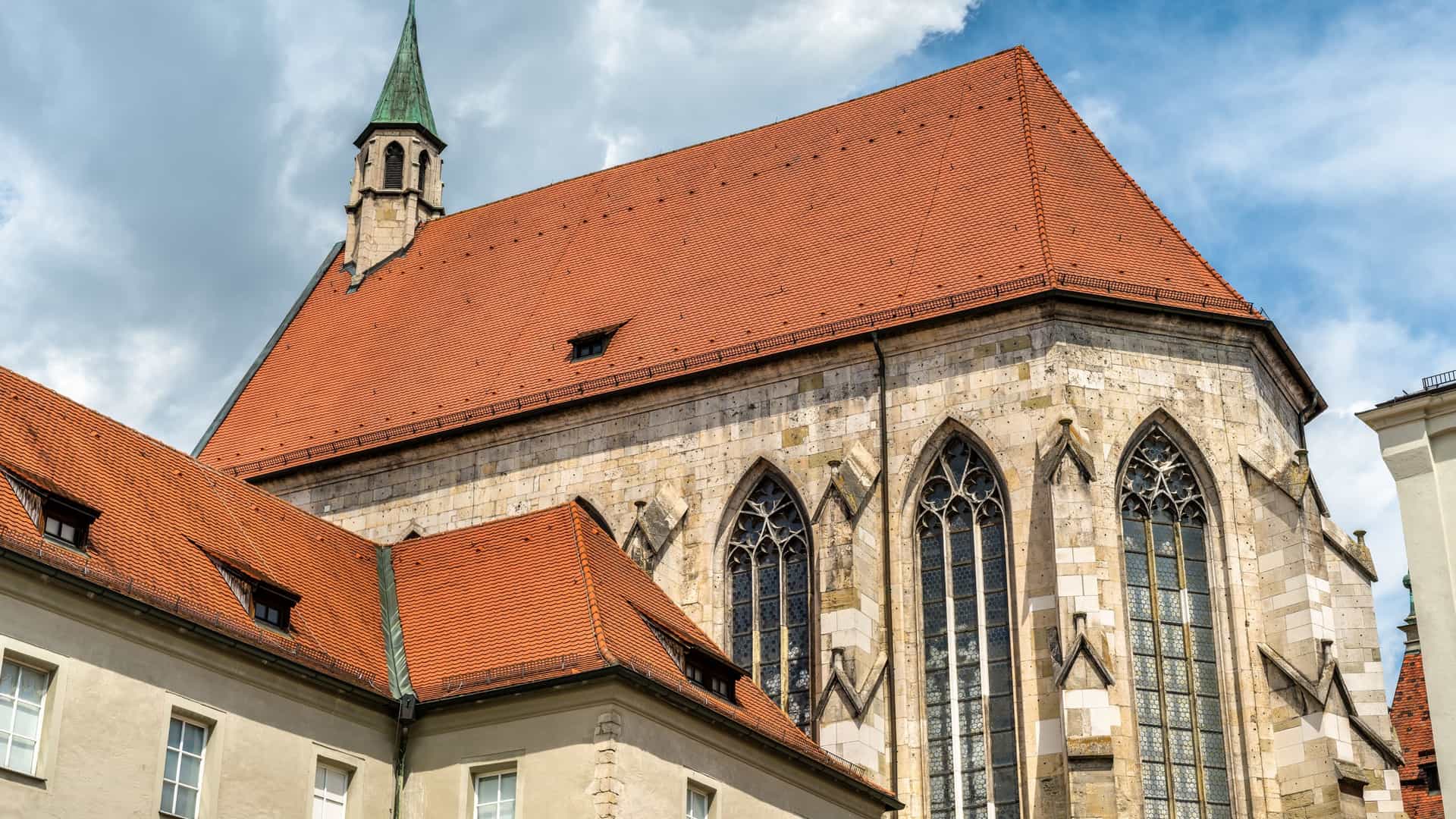 A detailed exterior view of the Historisches Museum Regensburg in Germany, which is housed in the former Minorite Church. The photo highlights the church's Gothic architecture, featuring large pointed arch windows, a terracotta tile roof, and an attached spire, next to a smaller, historic wing of the building.